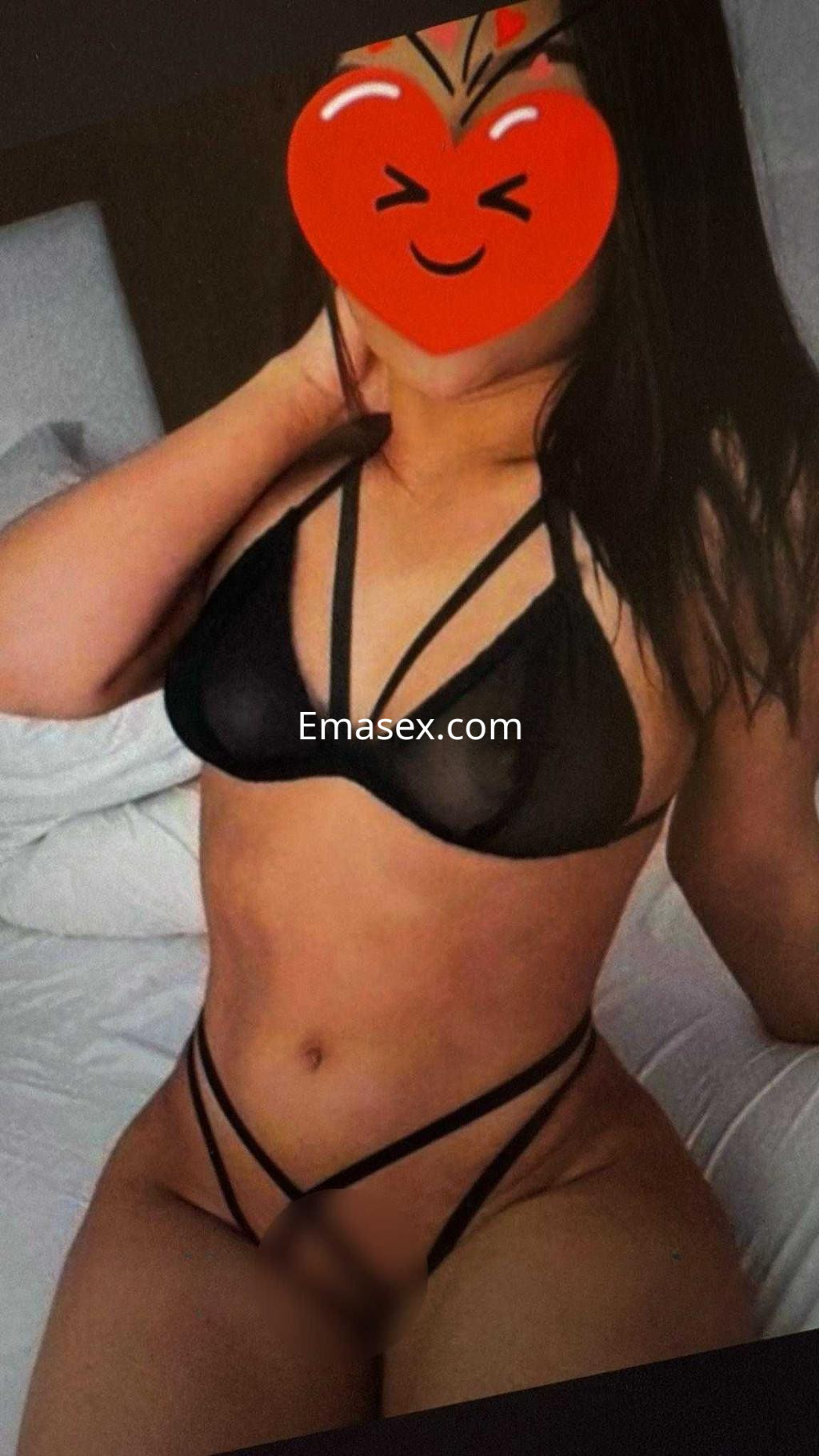 Puta en Alicante viste un conjunto de lencería negra con tiras, posando en una cama con fondo neutro.