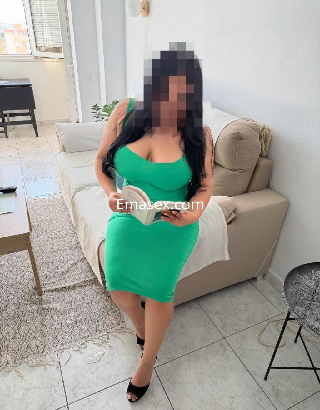 Escort en Barcelona, sentada en un sofá, viste un vestido verde ajustado y sostiene un libro.