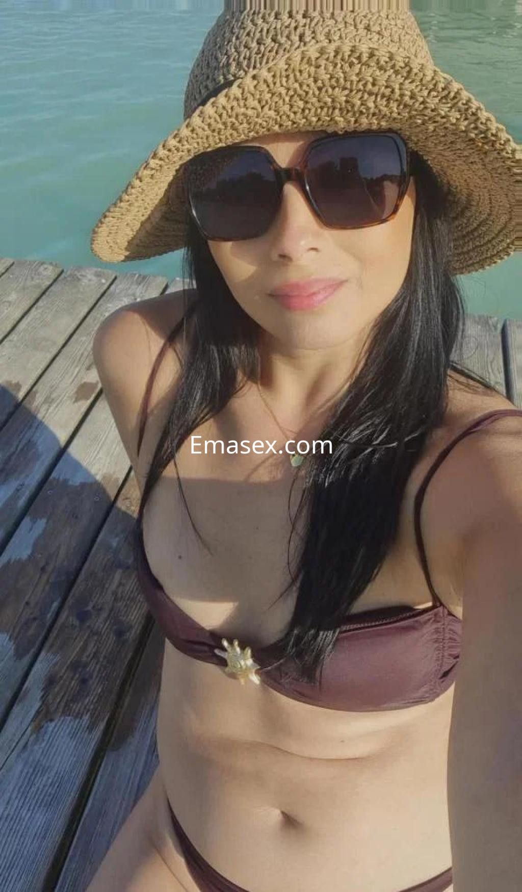 Puta en Barcelona luciendo un bikini marrón y un sombrero de paja, sonriendo frente a un entorno acuático.