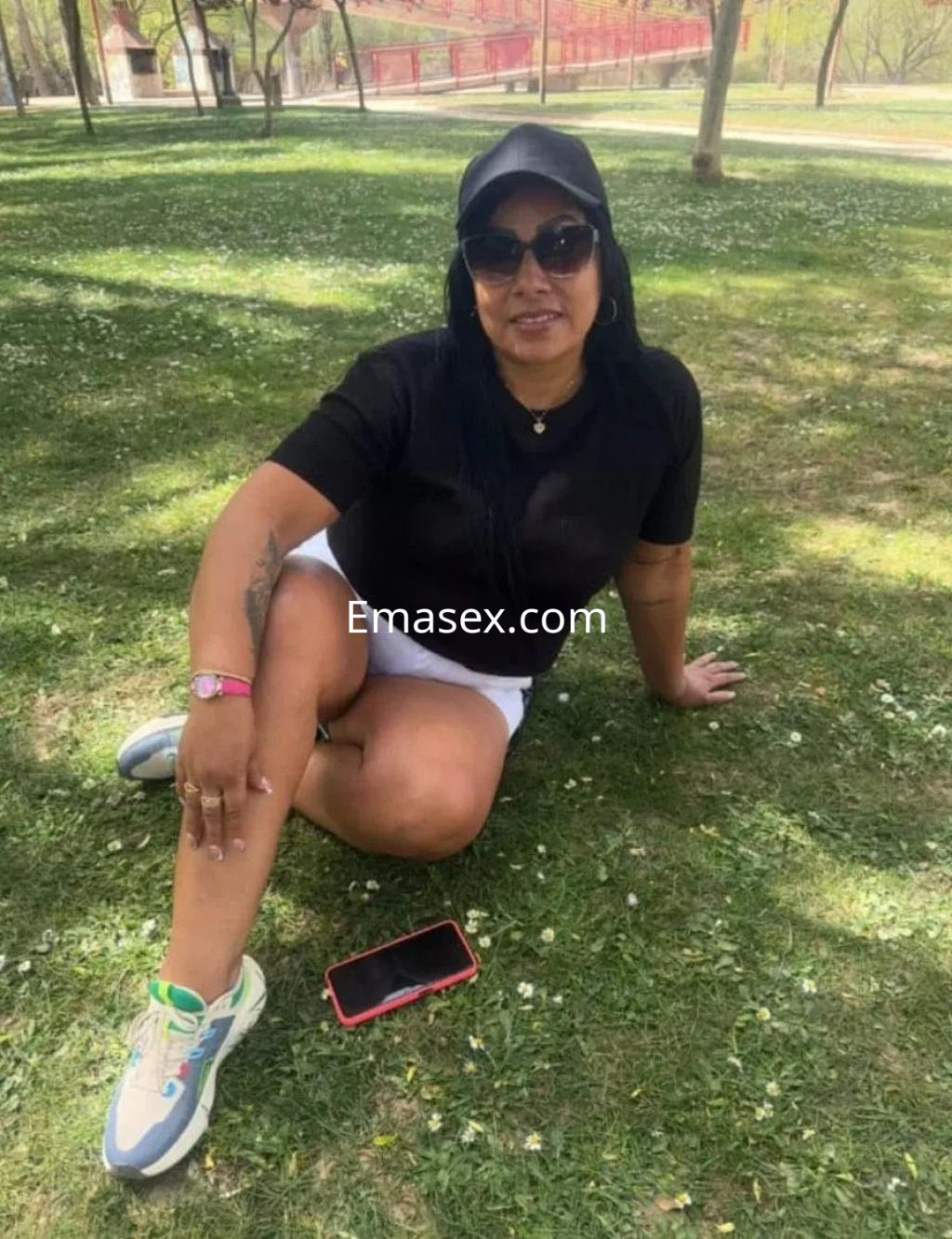 Escort en Burgos sentada en el césped, vistiendo camiseta negra y pantalones cortos, con gafas de sol y zapatillas deportivas.