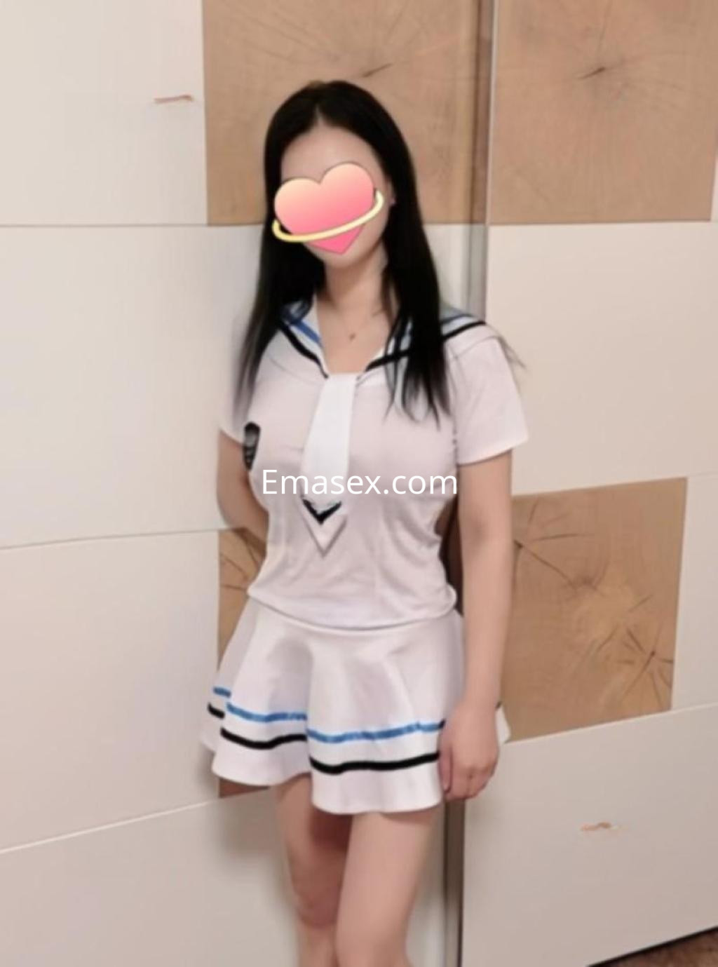 Escort en León con cabello largo y oscuro, vestida con uniforme escolar blanco y azul, posando de pie.