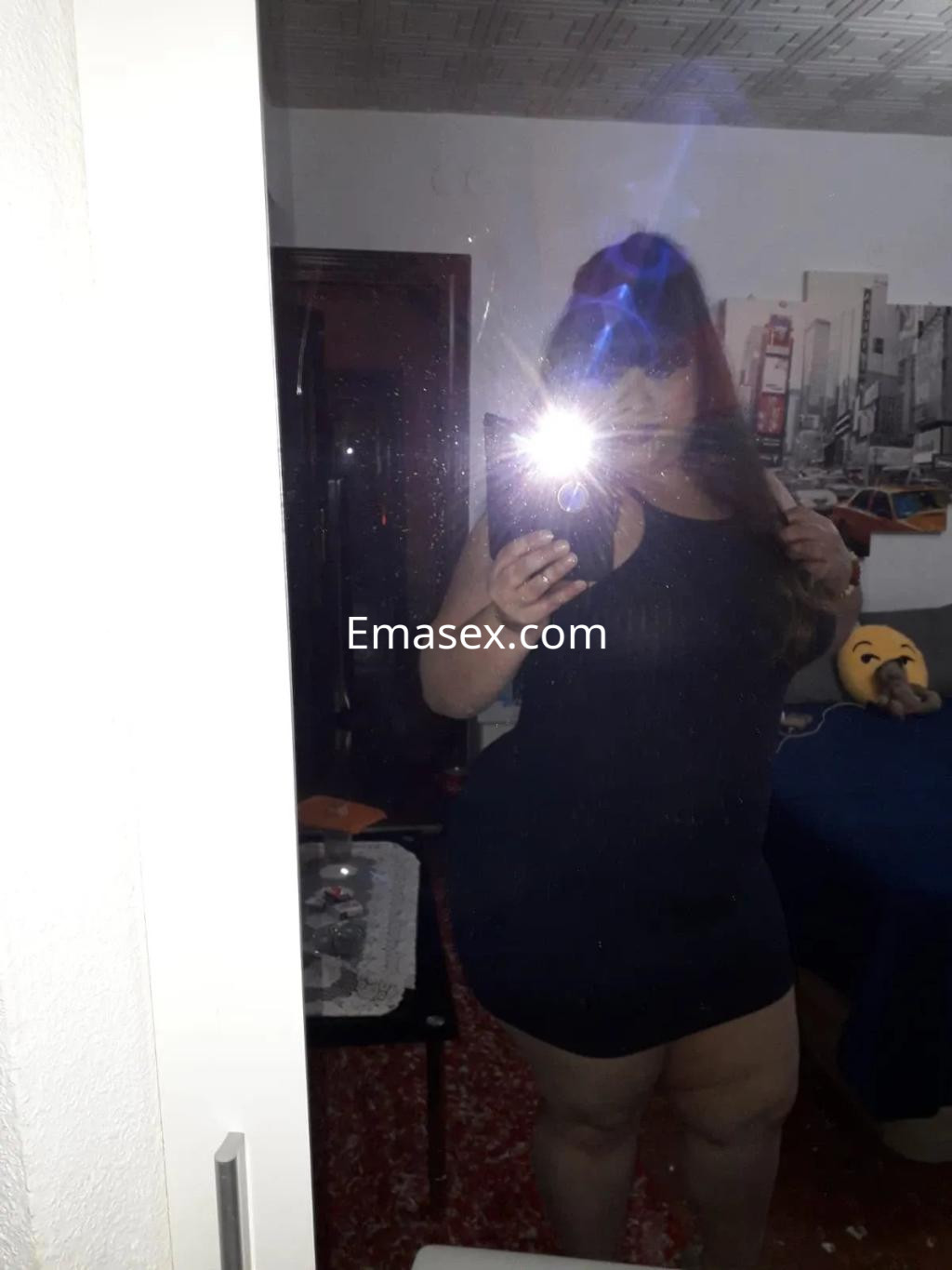 Puta en Sevilla posando frente a un espejo, con un vestido negro ajustado y cabello largo, en un ambiente interior.