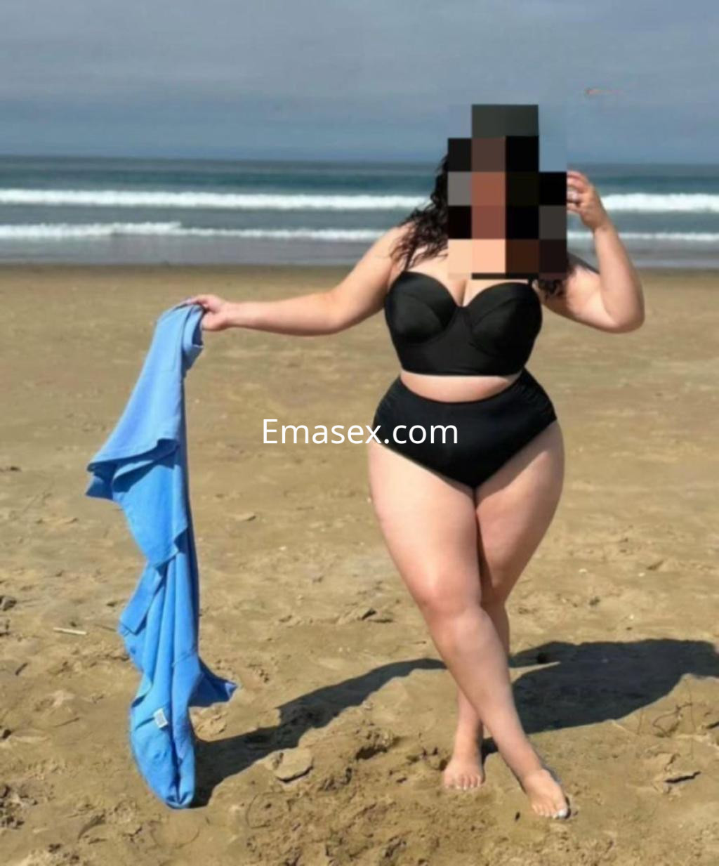 Escort en Tarragona con un bikini negro, posando en la playa y sosteniendo una toalla azul junto al mar.