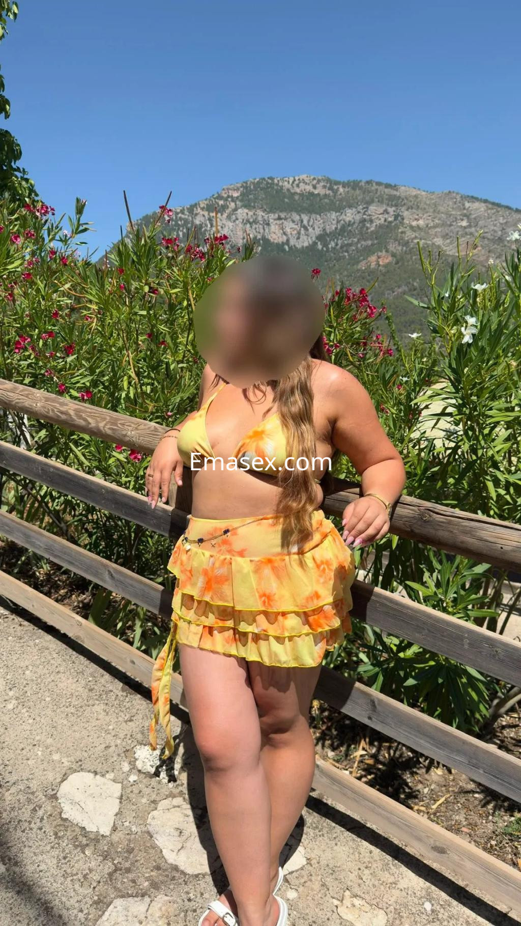 Escort en Valencia viste un conjunto amarillo con volantes, posando junto a una barandilla en un entorno natural.