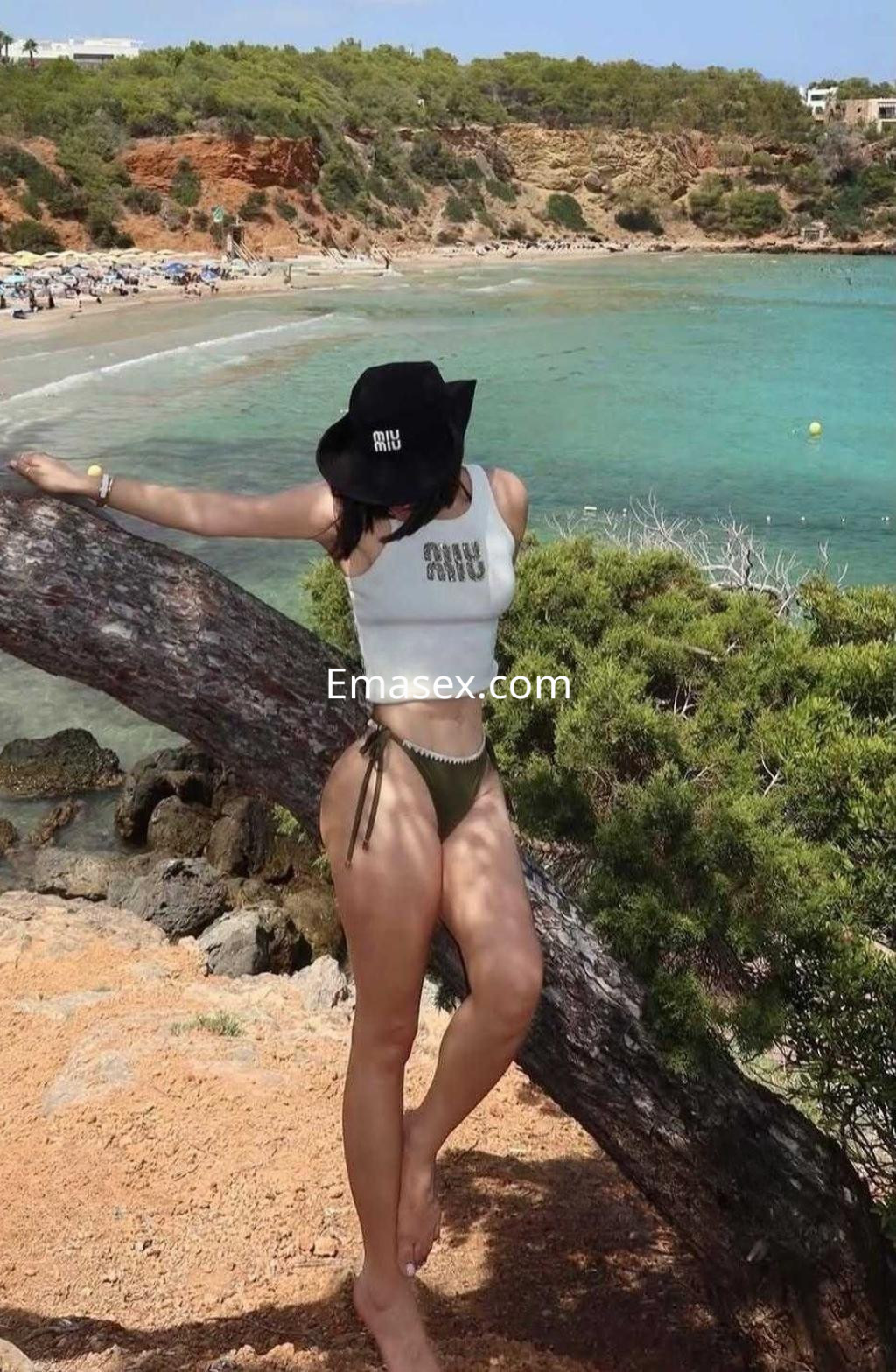 Escort en Tarragona, posando en la playa con un sombrero negro y un conjunto de baño verde, rodeada de naturaleza.