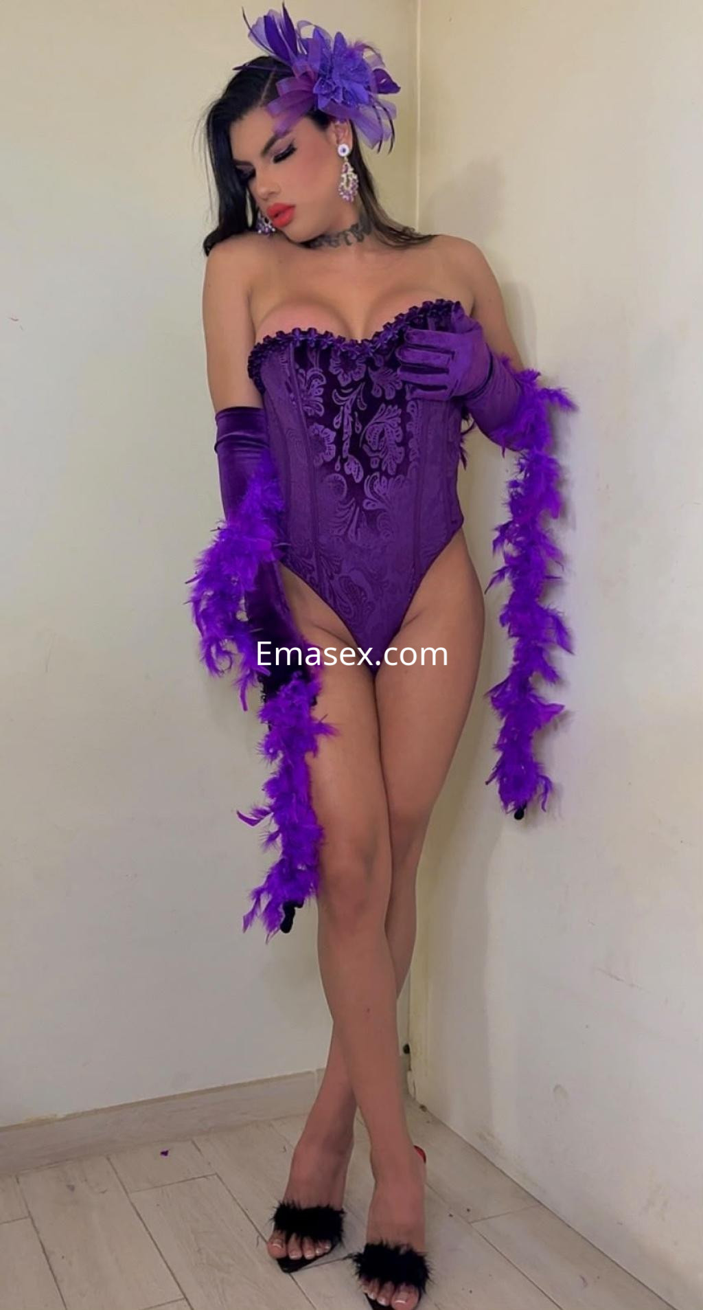 Escort trans en Irún luciendo un corsé morado con detalles, guantes largos y plumas, posando con elegancia.