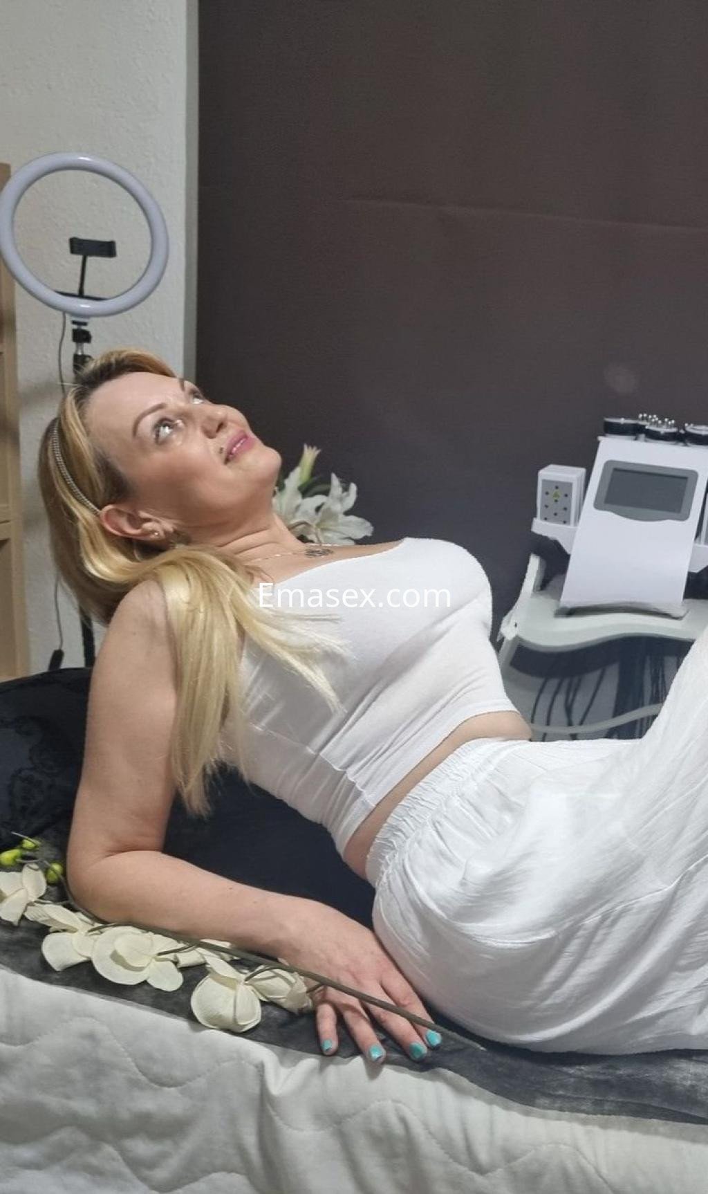 La puta está recostada sobre una cama con sábanas claras, usando una blusa blanca y un pantalón ligero en Málaga.