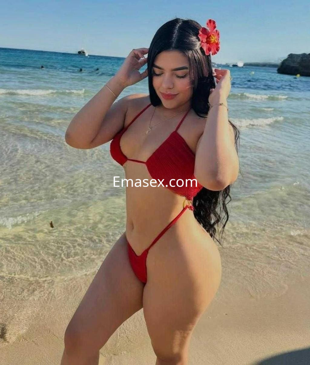 Puta en Vigo luciendo un bikini rojo en la playa, con cabello largo y flor en la cabeza, disfrutando del sol.