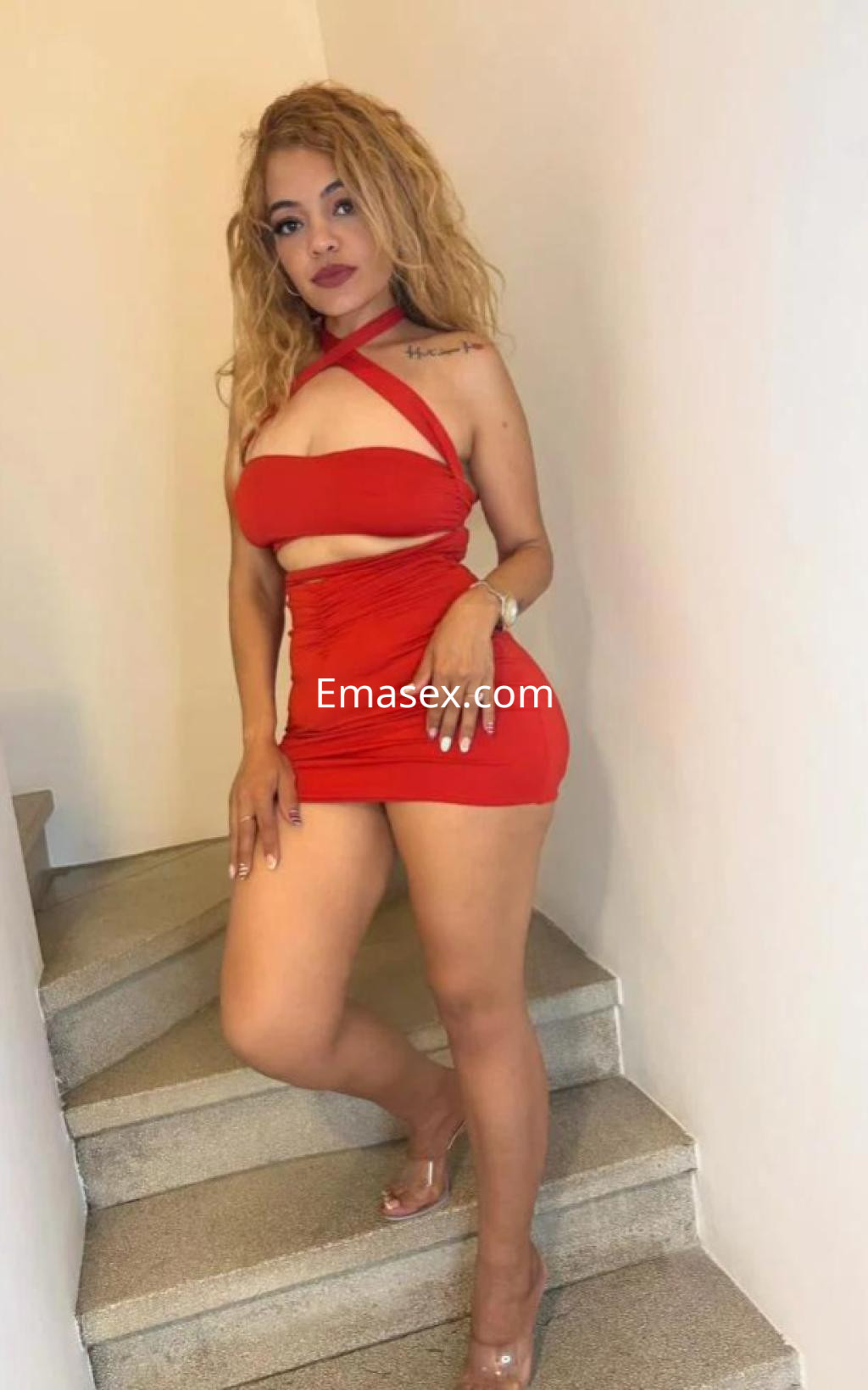 Escort en Cádiz, luciendo un vestido rojo ajustado y tacones, posando con confianza en una escalera.
