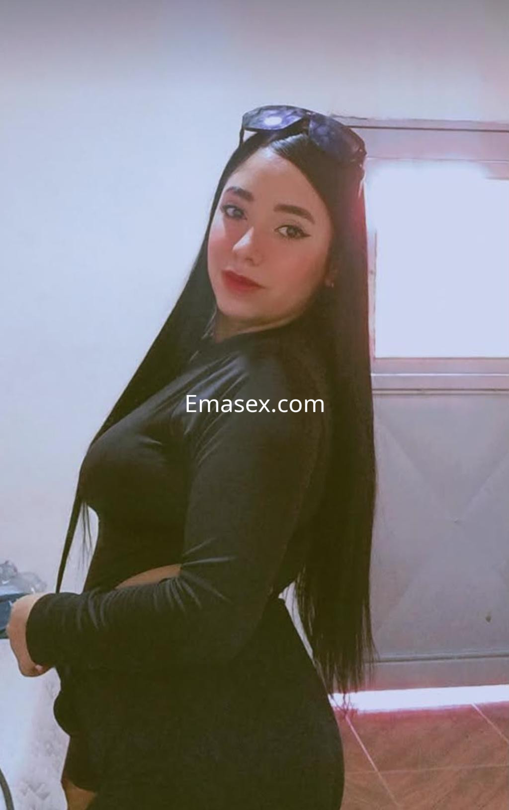 Escort en Bilbao, con cabello largo y lacio, viste una blusa negra ajustada y posa de perfil junto a una ventana.