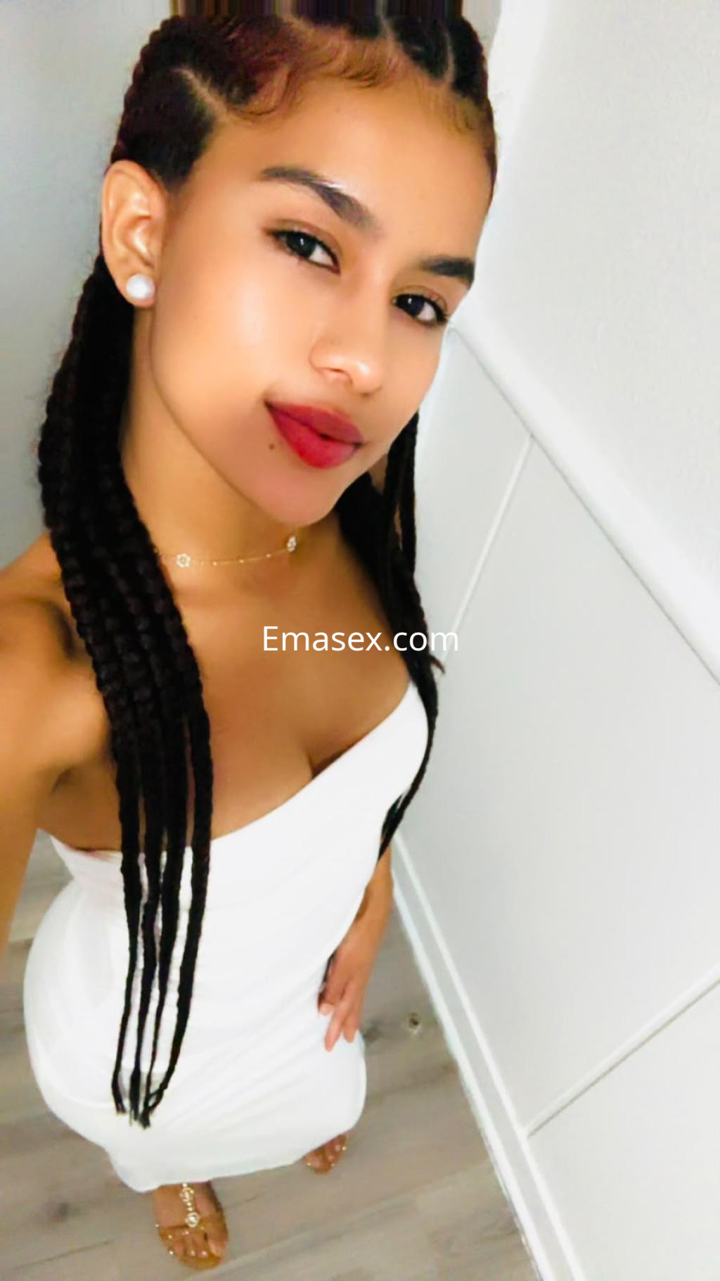 Escort en Alicante con cabello trenzado, luciendo un vestido blanco ajustado y joyas en un ambiente interior.