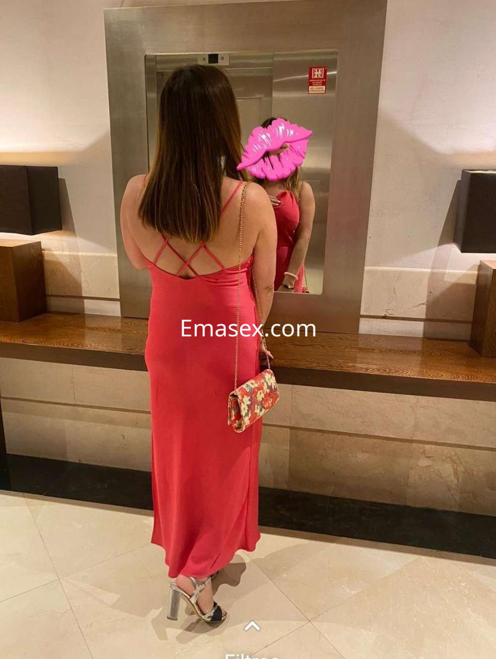 La puta en Albacete lleva un vestido largo fucsia y posa de espaldas frente a un espejo, luciendo un bolso floral.