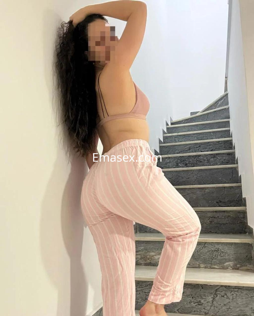 Escort en Alicante posando de perfil, con cabello rizado y ropa interior de color rosa y pantalones a rayas.