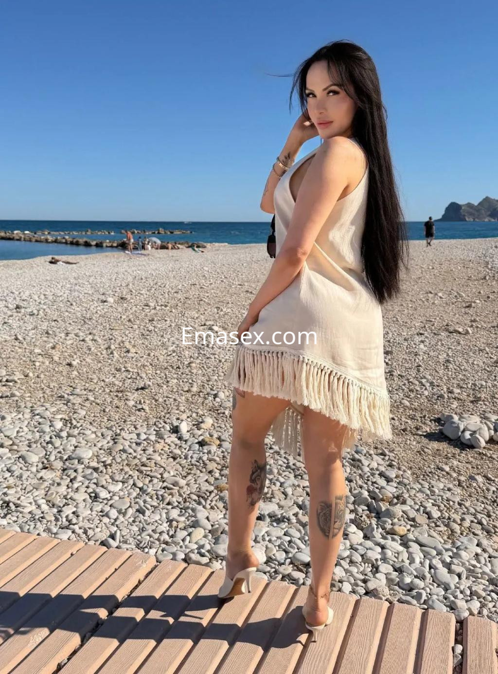 Puta de cabello largo y oscuro, viste un vestido ligero en la playa de Alicante, con tatuajes visibles en las piernas.