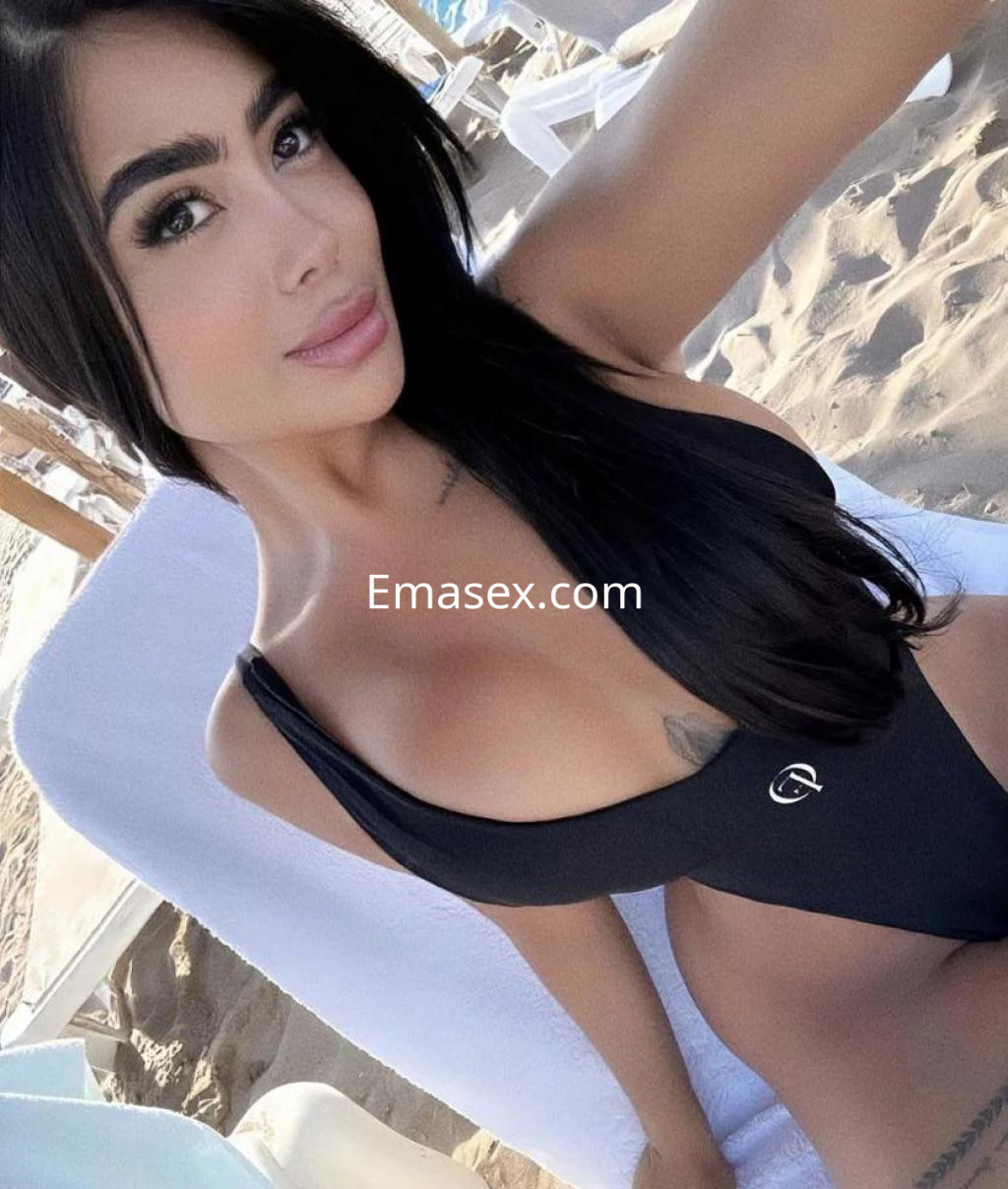 Escort en Palma de Mallorca posando en la playa, con un traje de baño negro y cabello largo y oscuro.