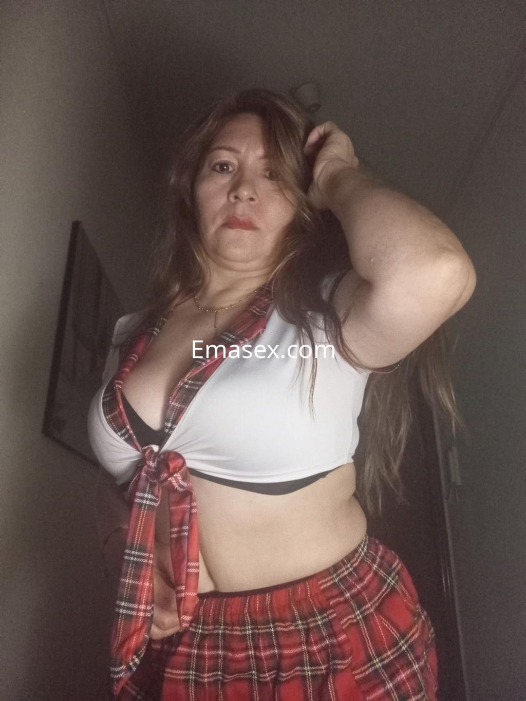 Escort en Barcelona con cabello largo y castaño, viste una blusa blanca atada y falda de cuadros rojos.