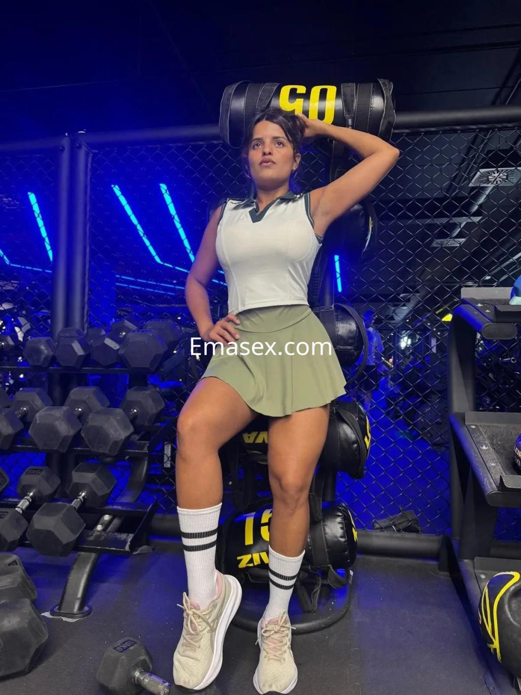 Escort en Cádiz, vistiendo un top deportivo blanco y una falda verde, posando en un gimnasio moderno.