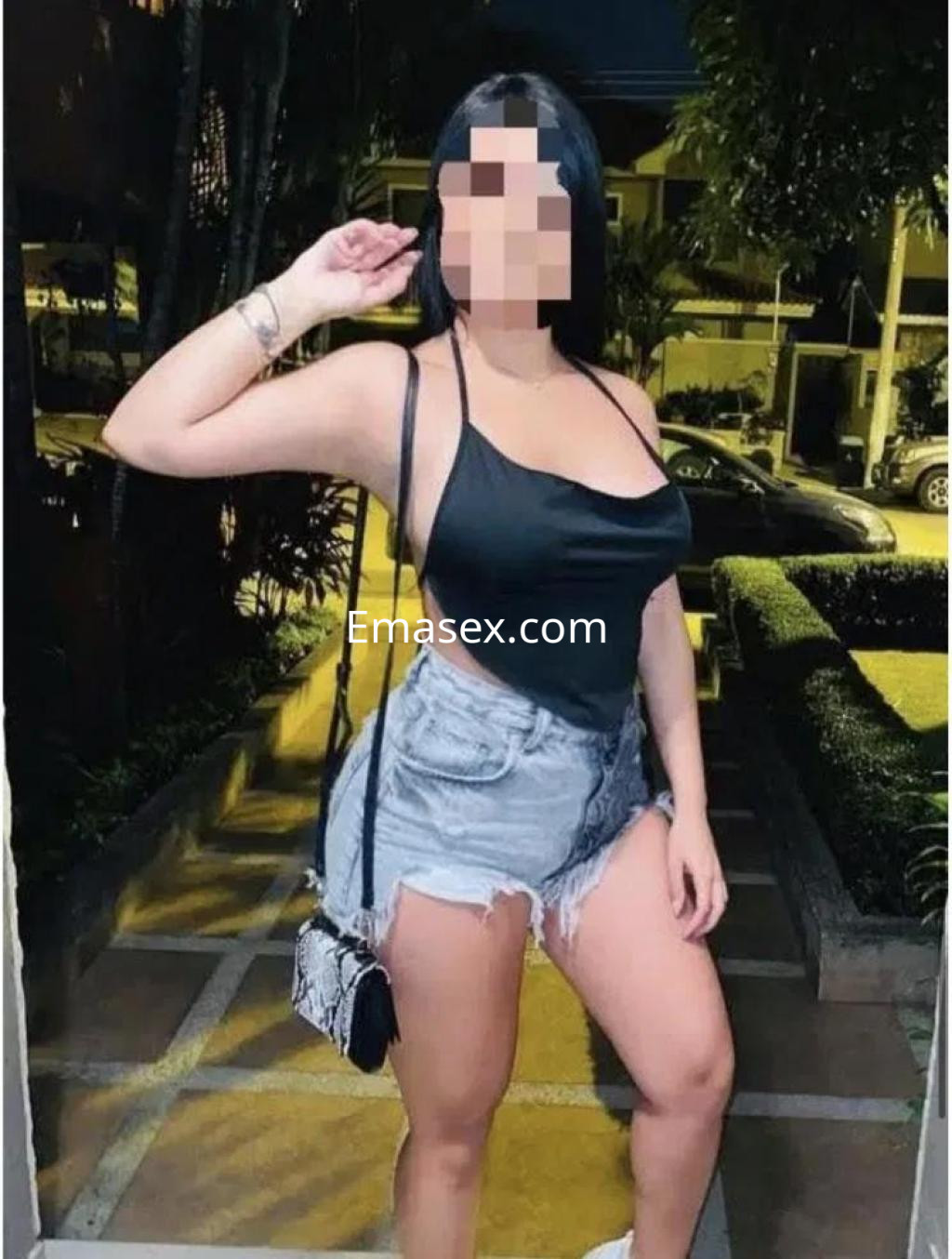 Escort en Castellón, posando con una blusa negra y shorts de mezclilla, con una bolsa de mano y un fondo urbano.