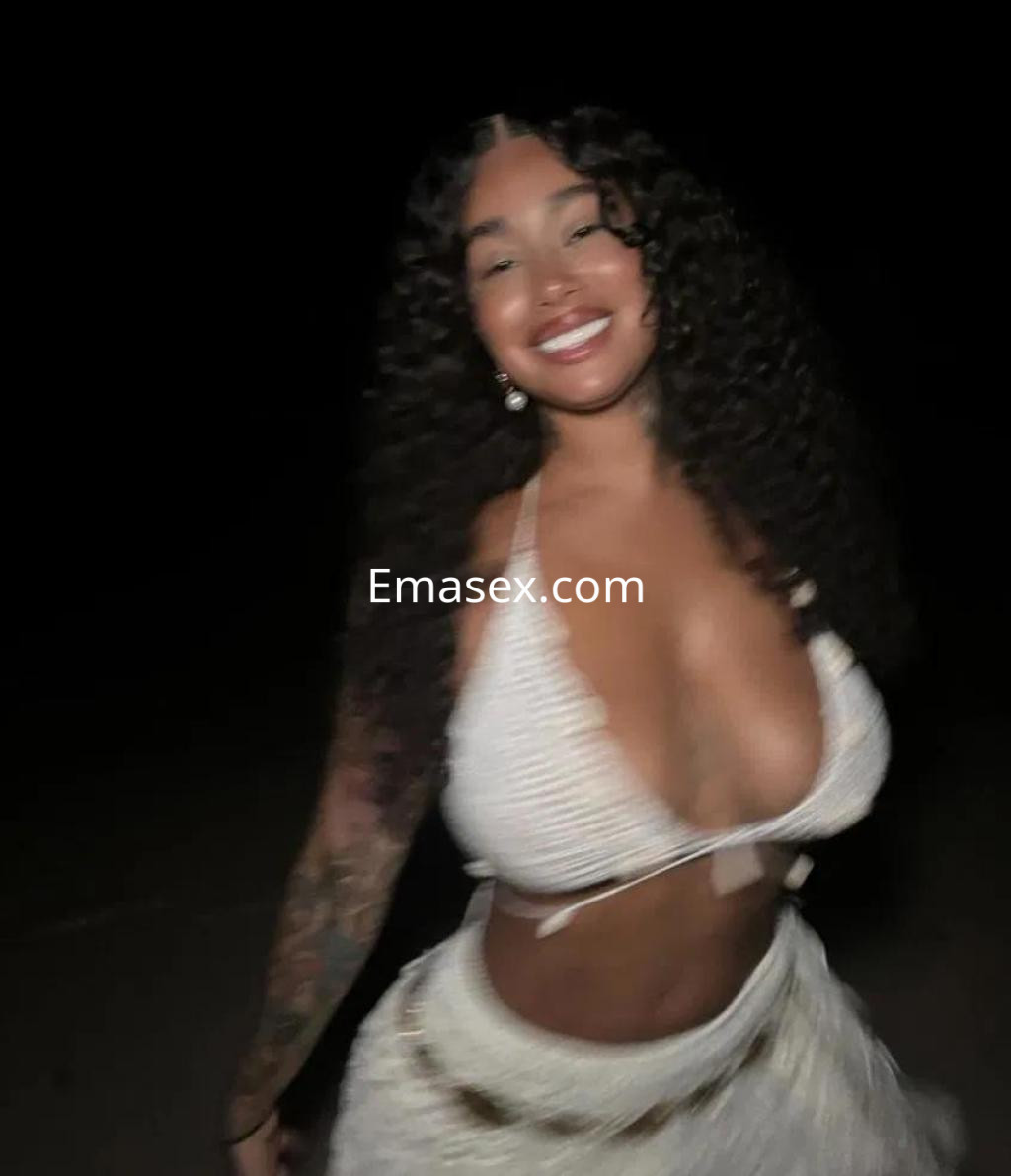 Escort en Coruña con cabello rizado y largo, vistiendo un top blanco y una falda de estilo bohemio en un entorno oscuro.