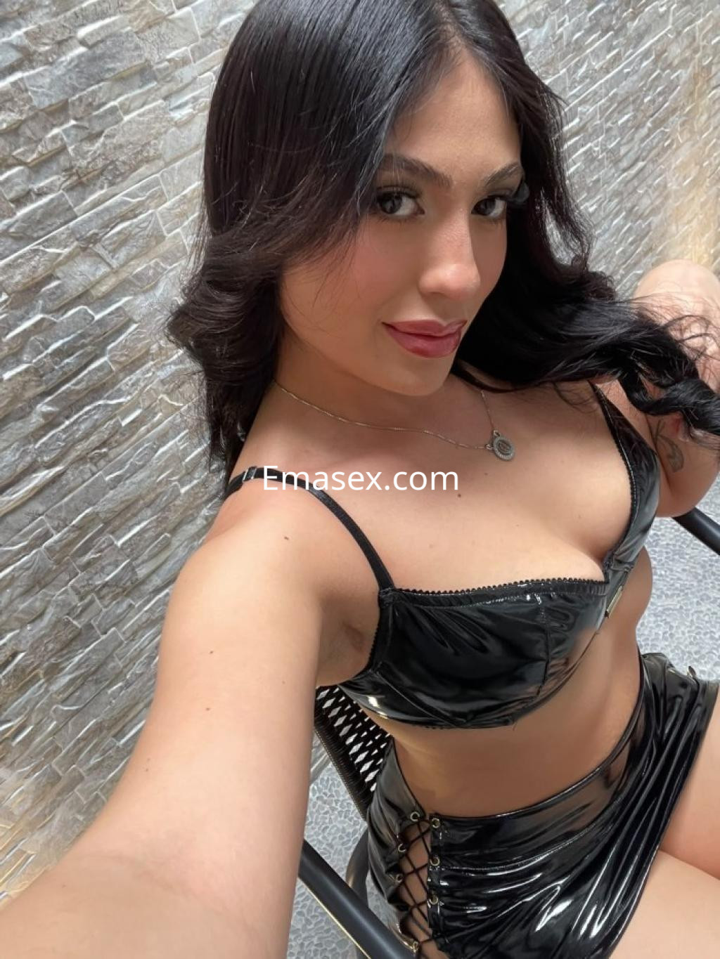 Escort en León con cabello oscuro y lacio, vistiendo un conjunto de lencería negra brillante en una pose coqueta.