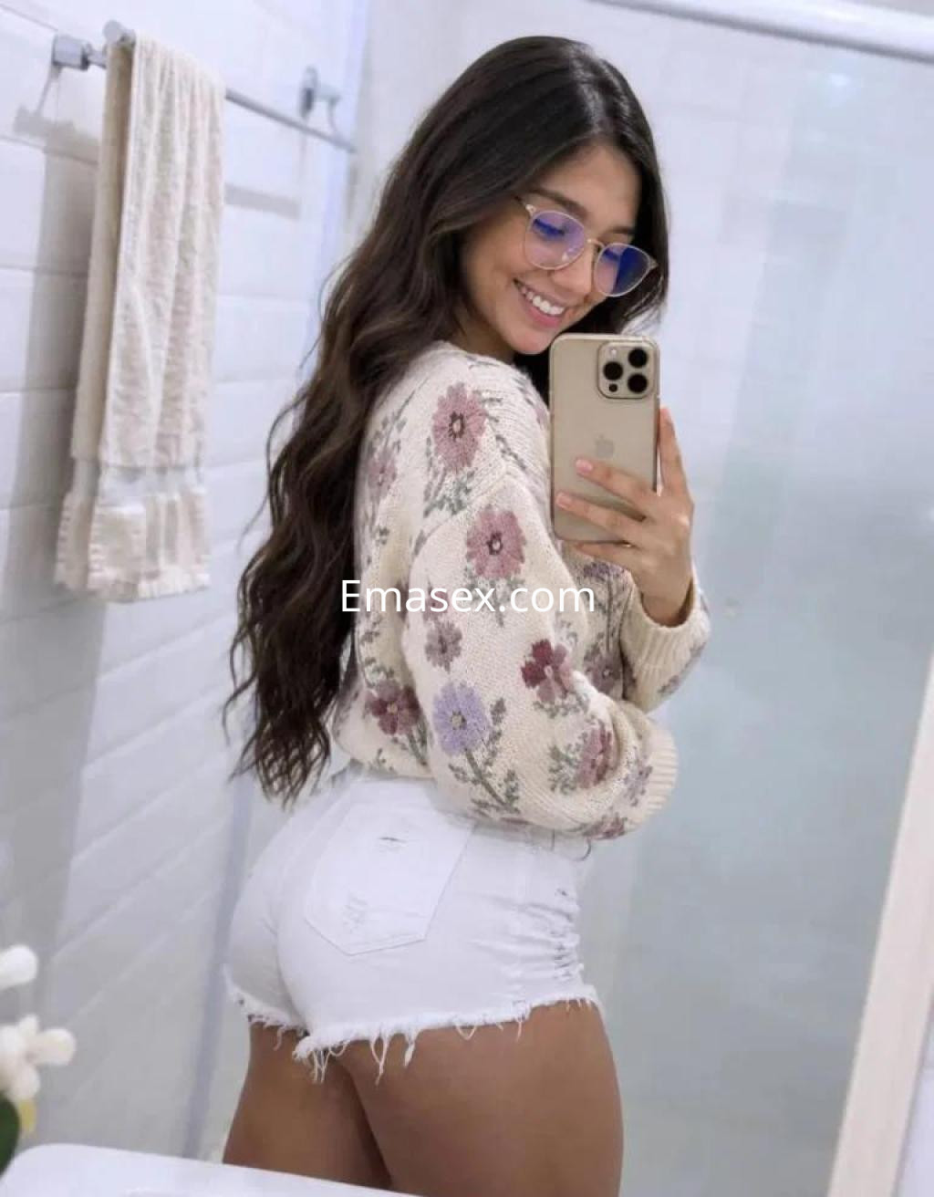 Escort en Lugo, con cabello largo y ondulado, viste un suéter claro con flores y shorts blancos, sonriendo en un espejo.