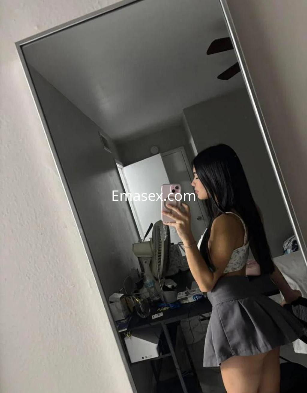 La puta luce un top blanco y una falda corta gris, posando frente a un espejo en un ambiente interior.