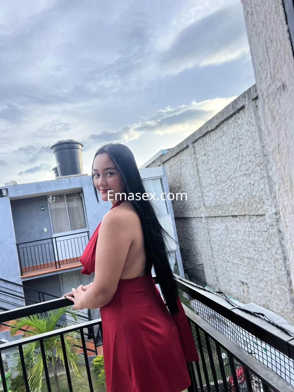 La escort en Vigo lleva un vestido rojo sin mangas y posa sonriente en un balcón con un fondo urbano.