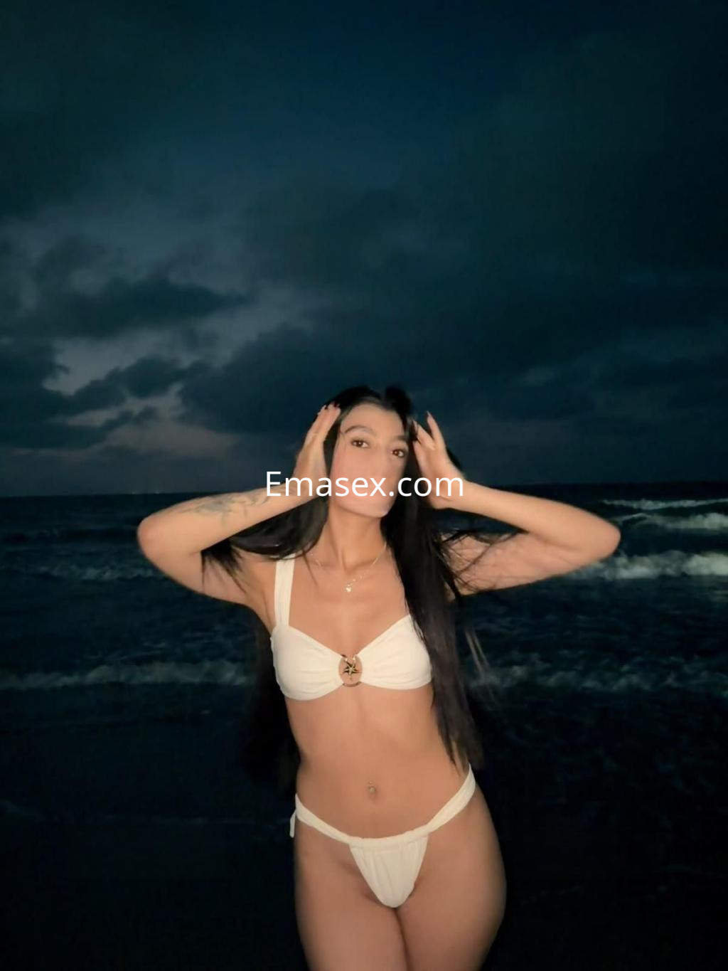 Escort en Tenerife vistiendo un bikini blanco, posando en la playa al atardecer con el mar de fondo.