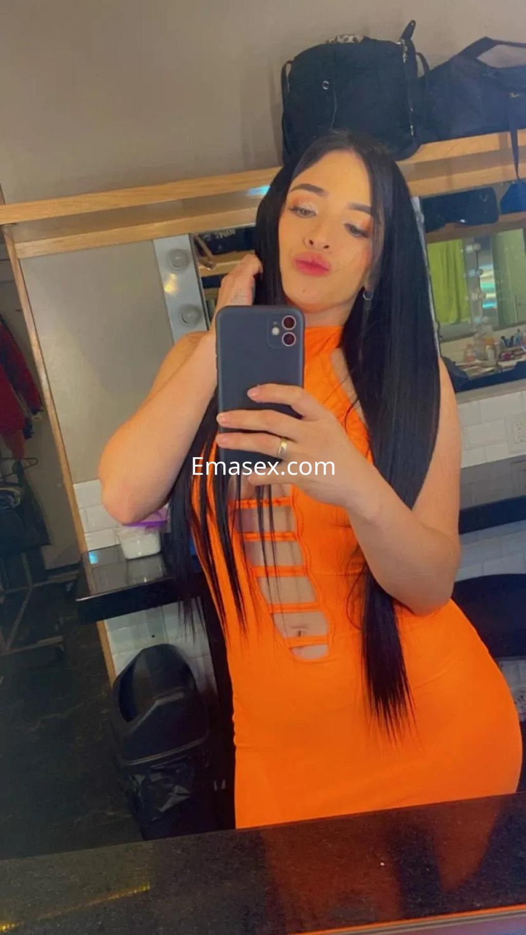 La puta posa frente al espejo con un vestido naranja ajustado, mostrando su largo cabello oscuro en Sevilla.