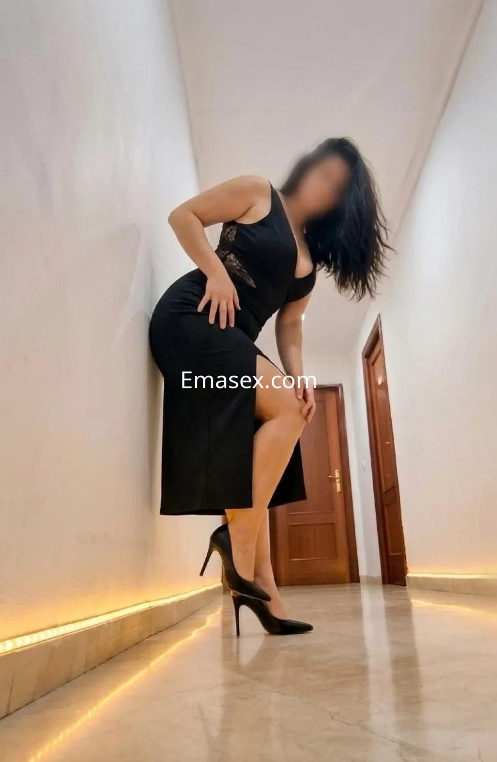 Puta en Sevilla, posando con un vestido negro ceñido y tacones altos, en un pasillo iluminado.
