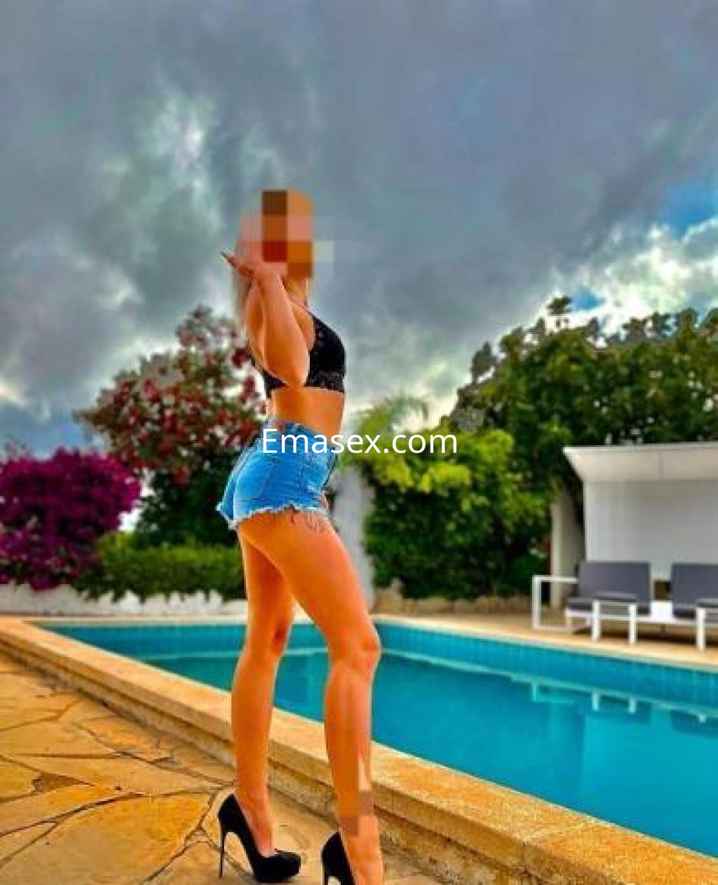 Escort en Tarragona posando junto a una piscina, viste shorts cortos y tacones altos, con fondo de vegetación.