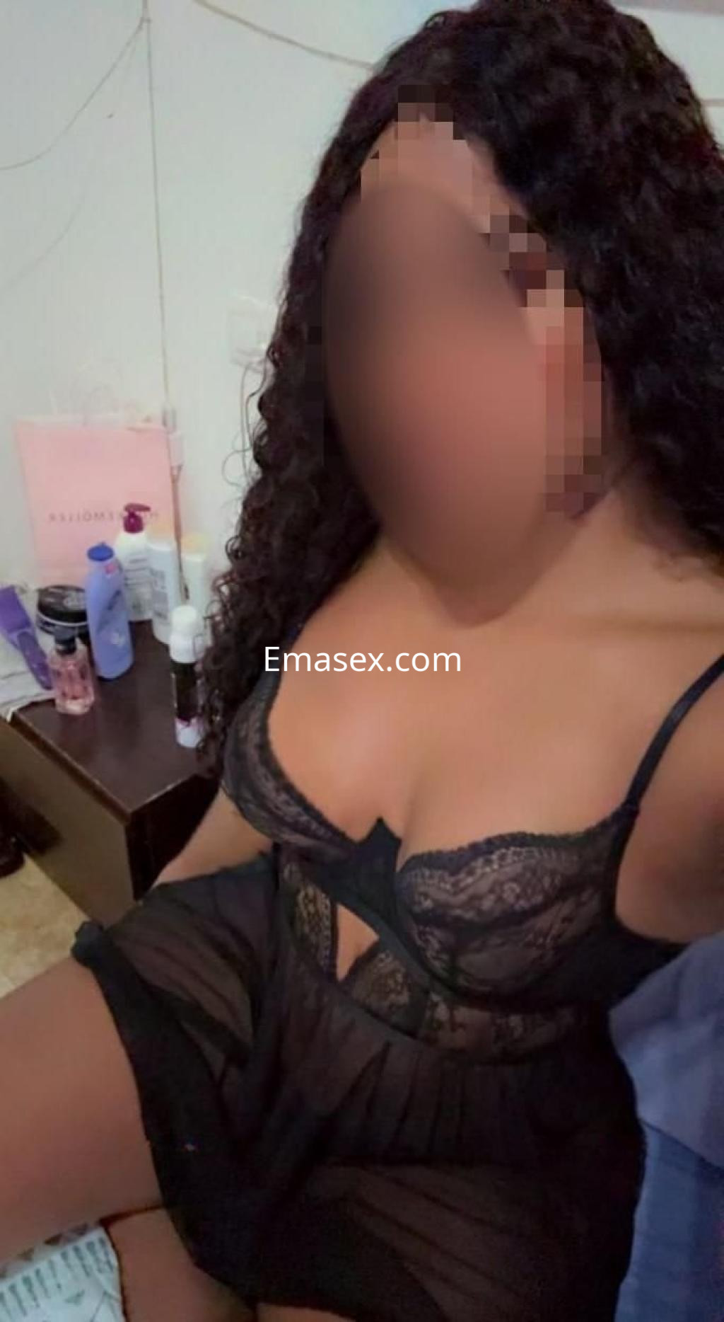 Puta en Tarragona viste una lencería negra con encaje, sentada en una cama con productos de belleza alrededor.