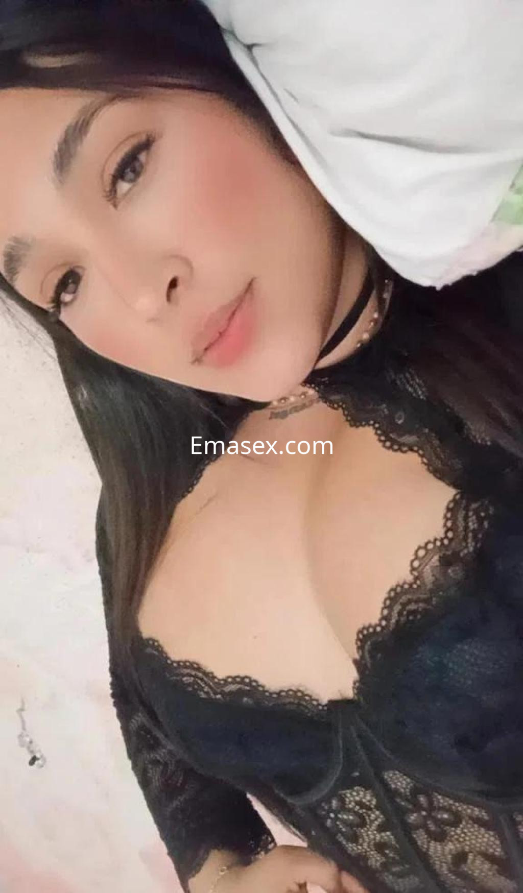 Escort en Toledo, posando de lado con un corsé negro de encaje y un collar, sobre una cama con sábanas claras.