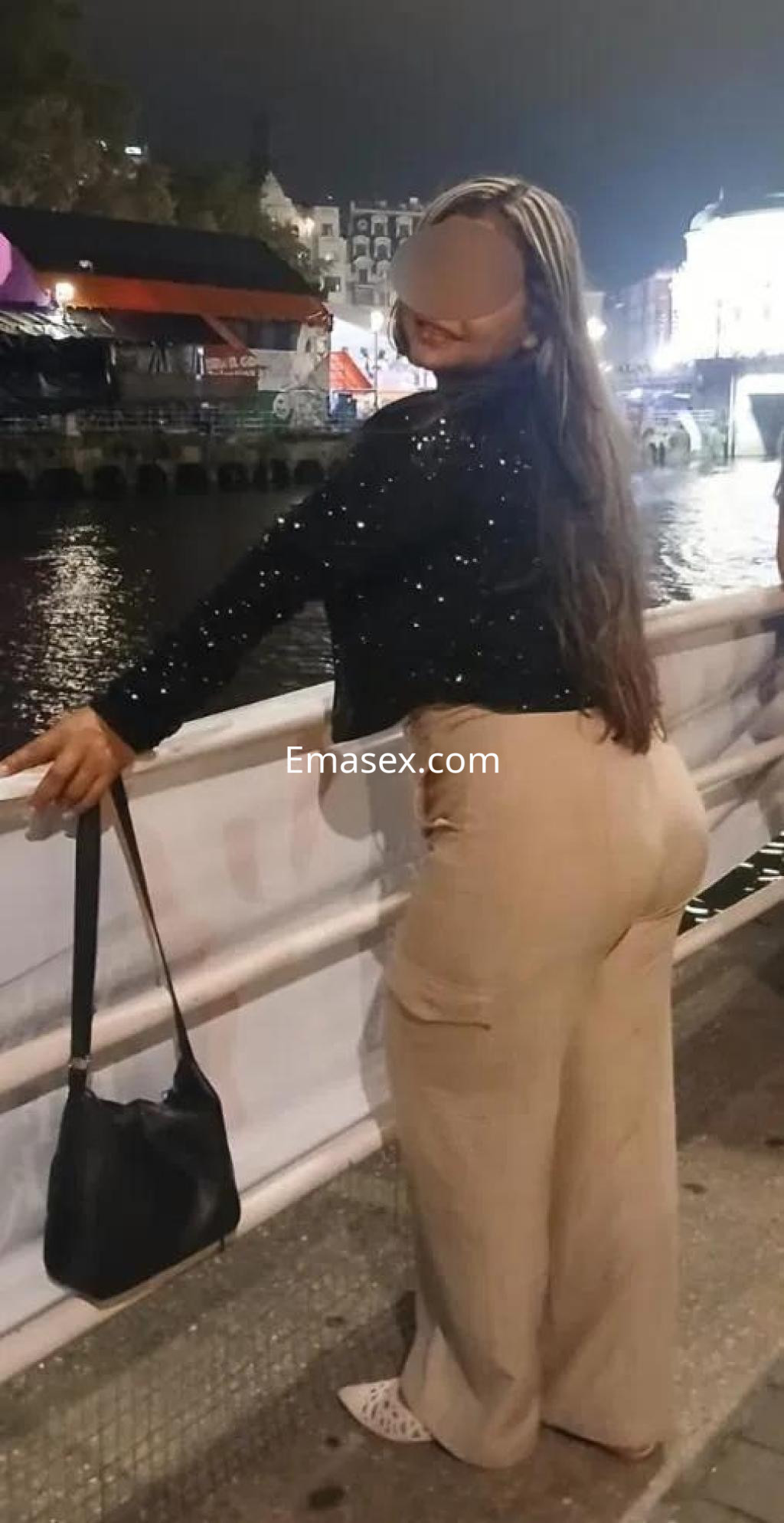 Escort de Bilbao con cabello largo y lacio, vistiendo blusa brillante y pantalones claros, posando junto al agua.