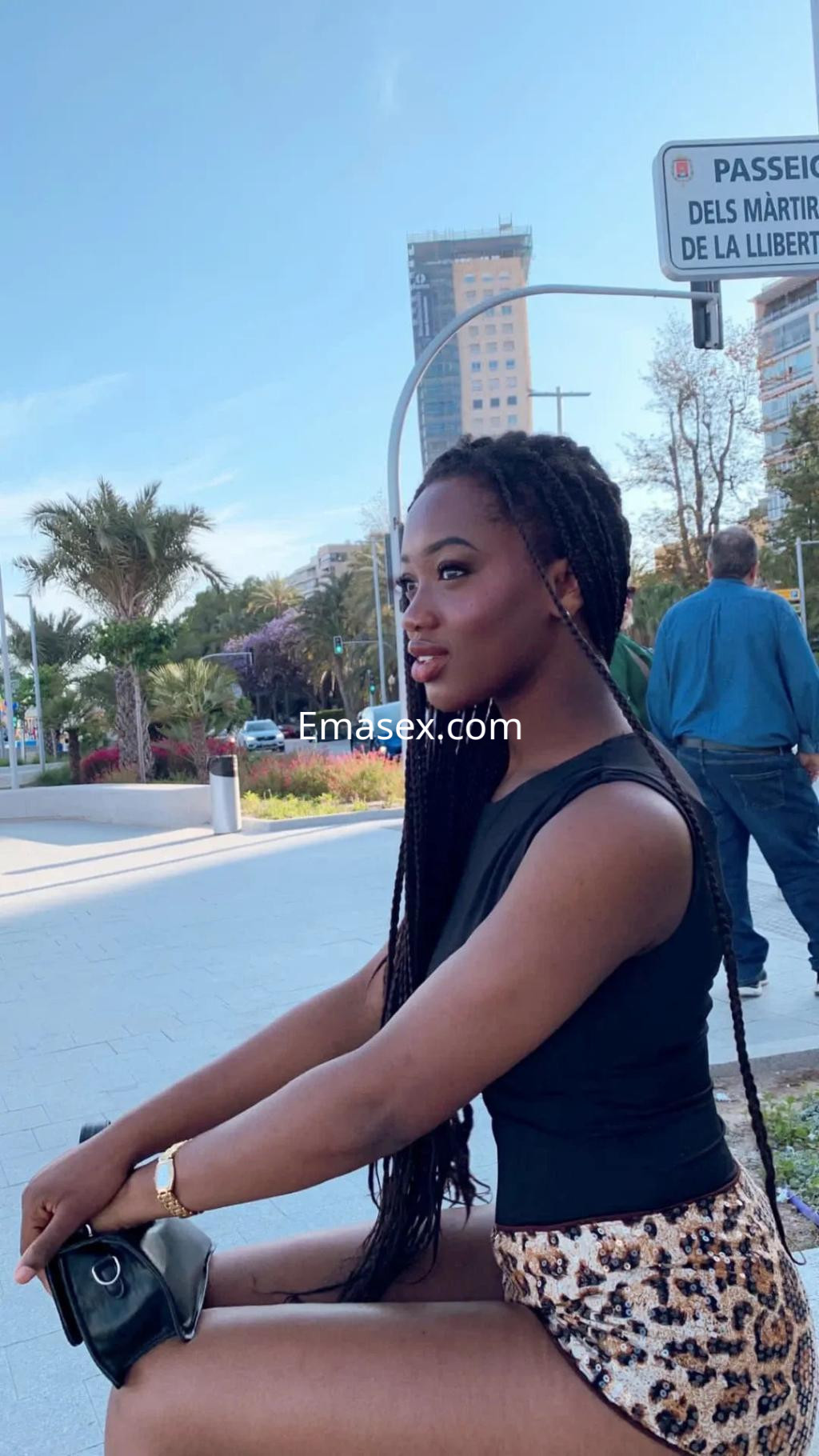 Escort en Alicante sentada al aire libre, vistiendo un top negro y una falda de leopardo, con trenzas largas.