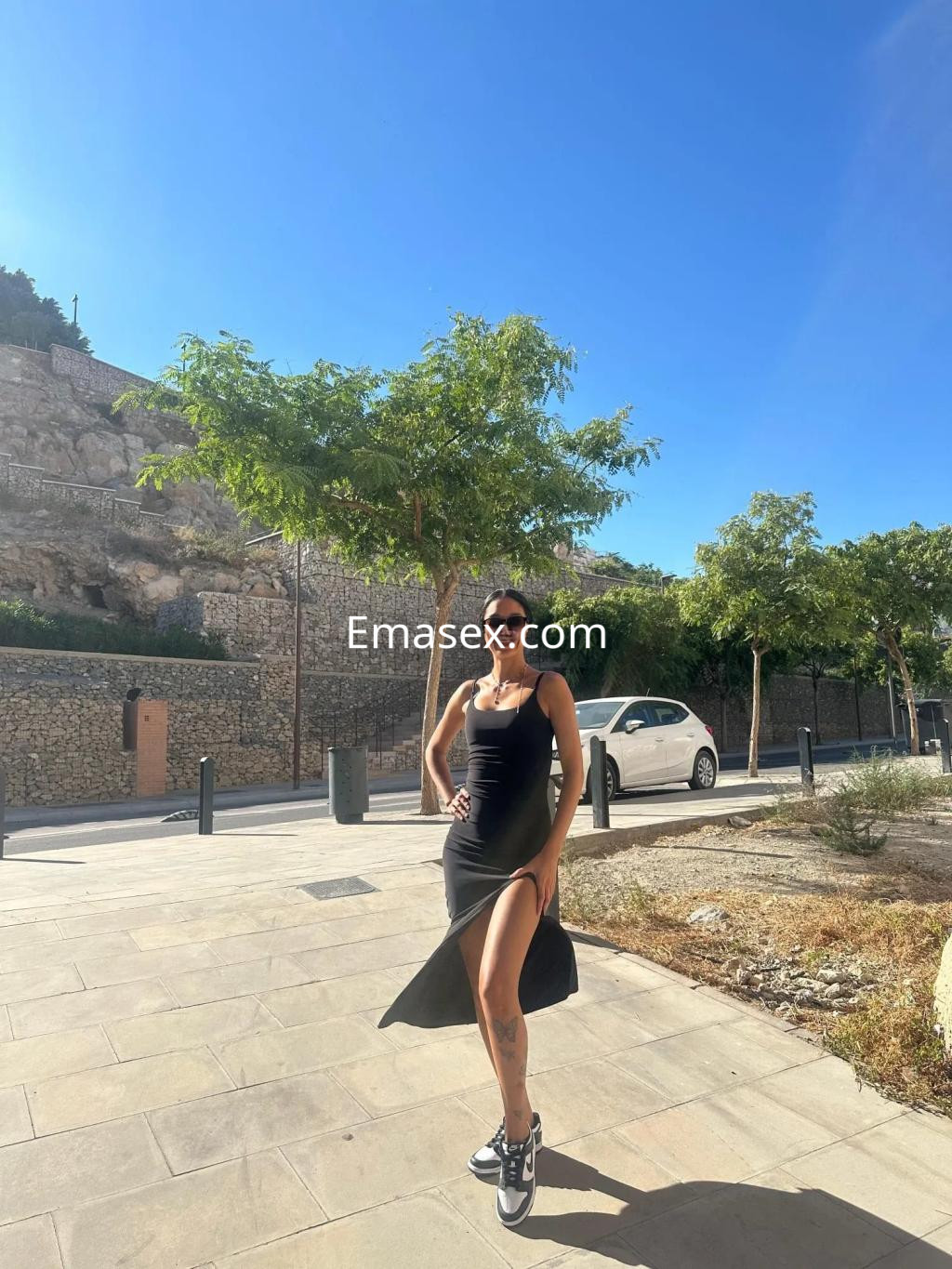 Escort en Alicante, con vestido negro ajustado y abertura, posando al aire libre bajo un cielo despejado.