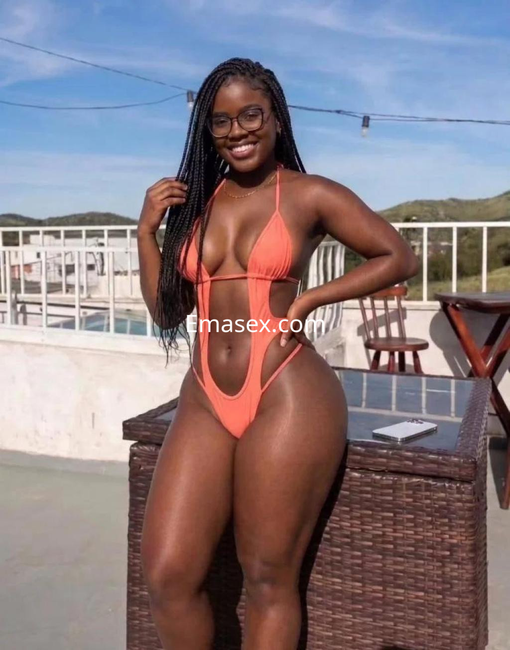 Puta en Badajoz luce un bikini naranja, con trenzas largas y gafas, sonriendo en un ambiente al aire libre.