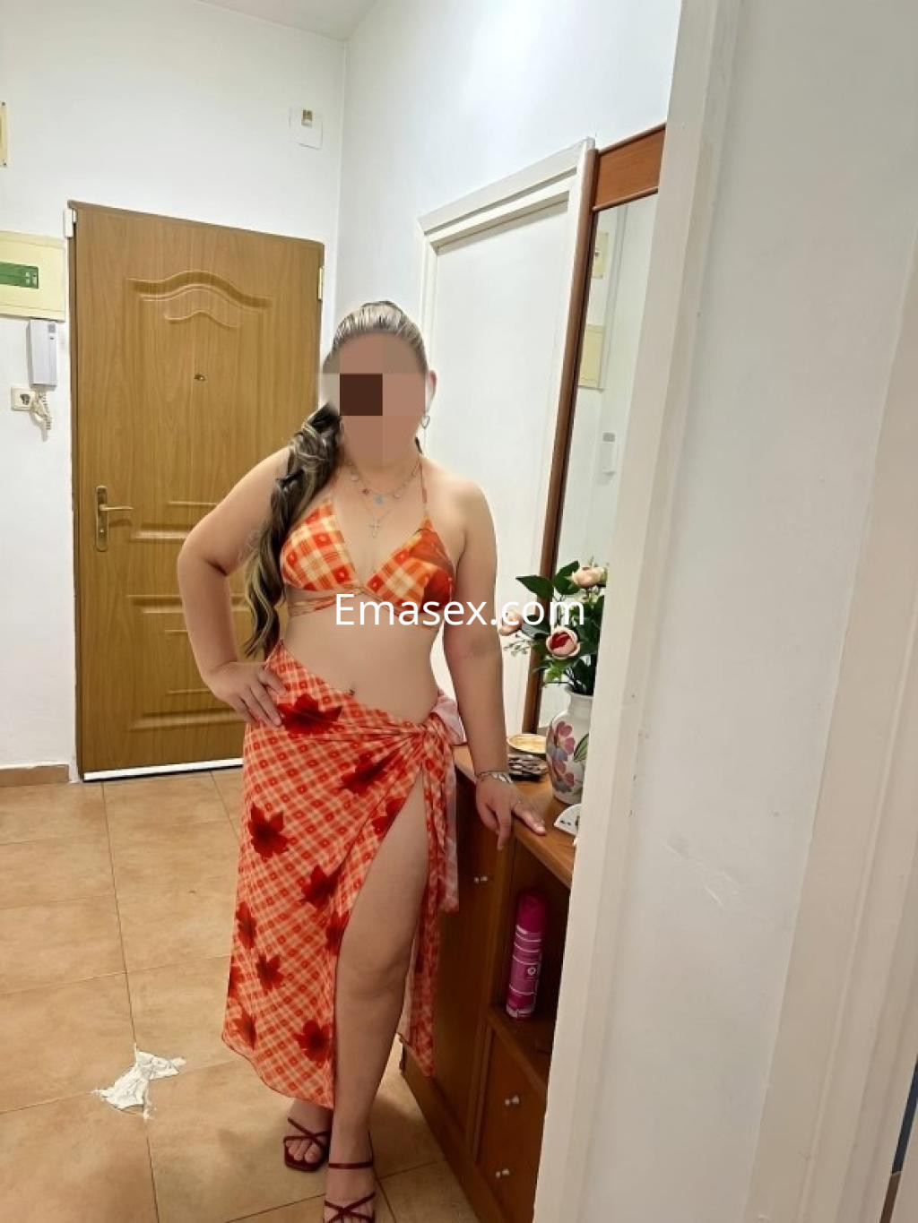 La puta en Santiago viste un conjunto de bikini naranja con un pareo a juego, posando en un entorno interior.