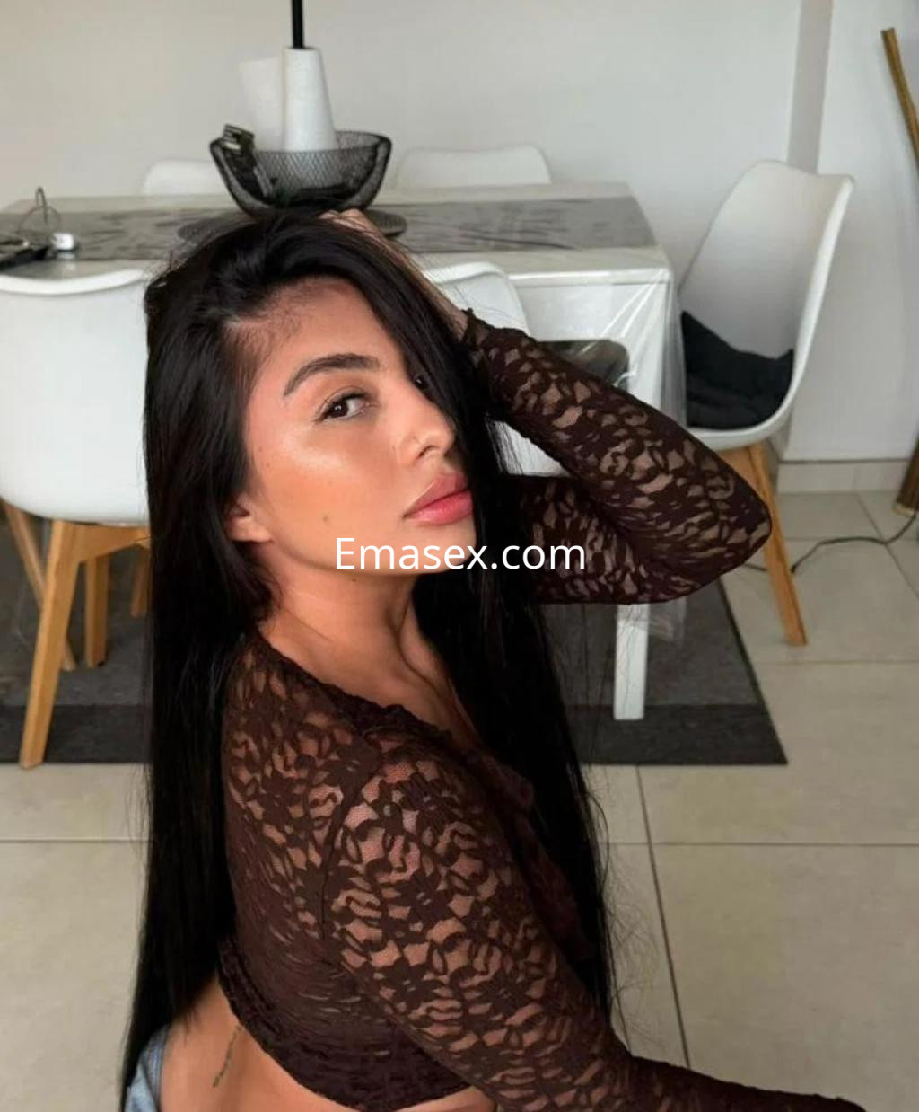 Puta de cabello largo y oscuro, viste una blusa de encaje marrón, posando en un interior moderno en Madrid.