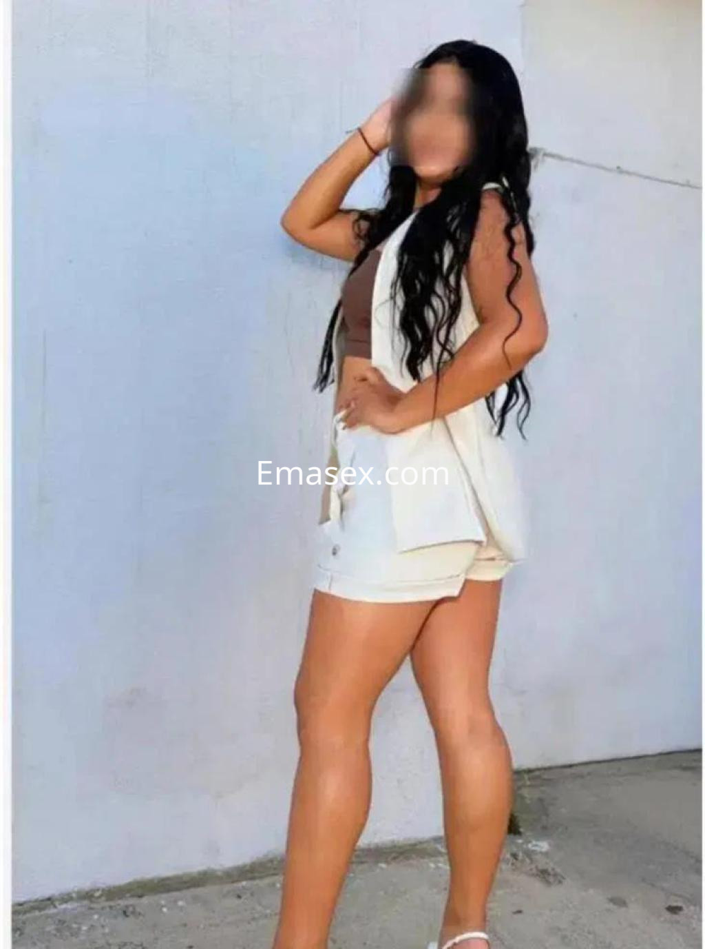 Escort en Madrid con cabello largo y rizado, vistiendo un conjunto claro de blusa y pantalones cortos, posando lateralmente.