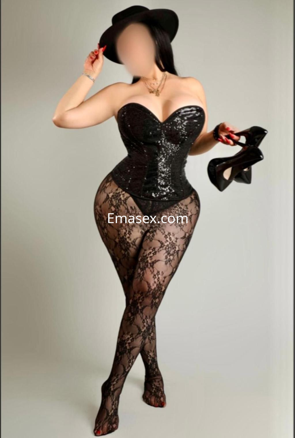 Escort en Madrid luciendo un corsé negro brillante y medias de encaje, posando con un sombrero y tacones.