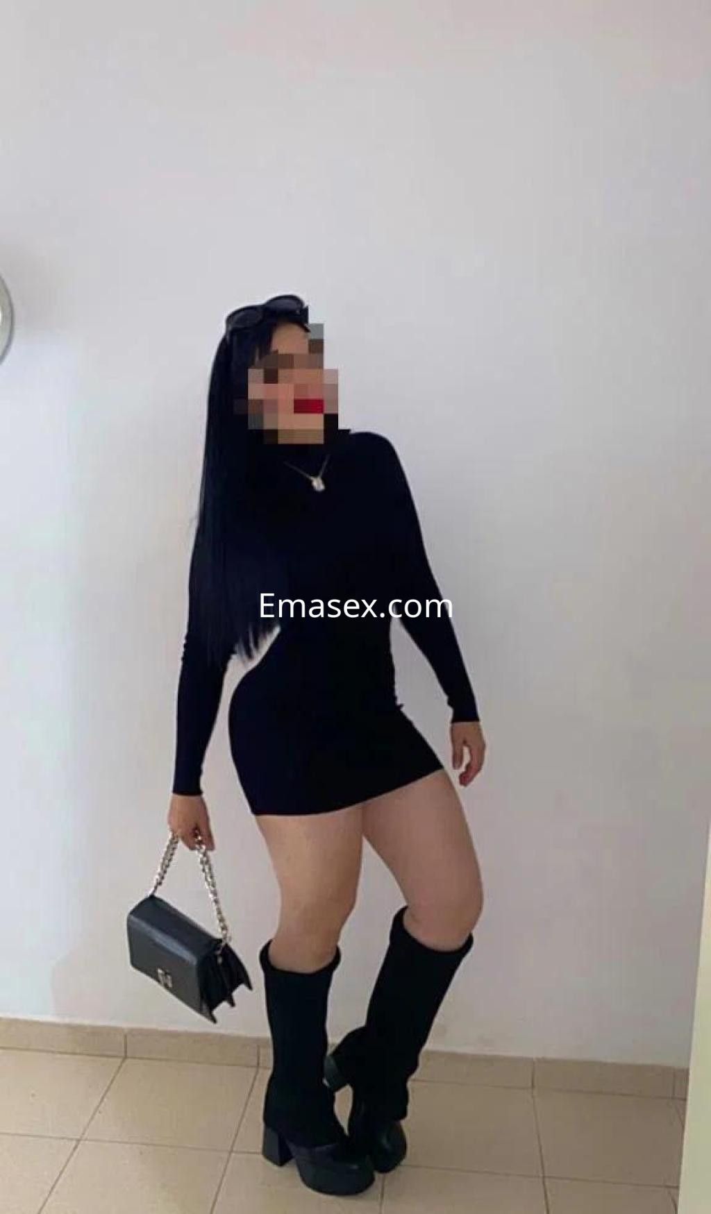 Puta posando en Ourense, viste un vestido negro ajustado y botas altas, con un bolso de mano a juego.