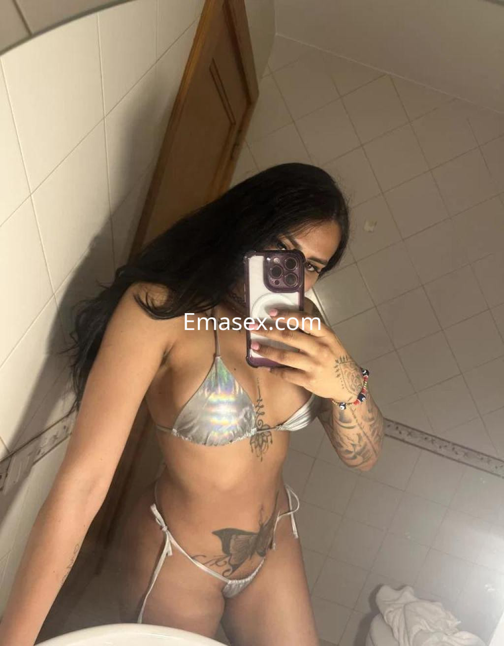 La puta luce un bikini plateado en un baño, con el cabello largo y oscuro, mostrando un tatuaje en el torso.
