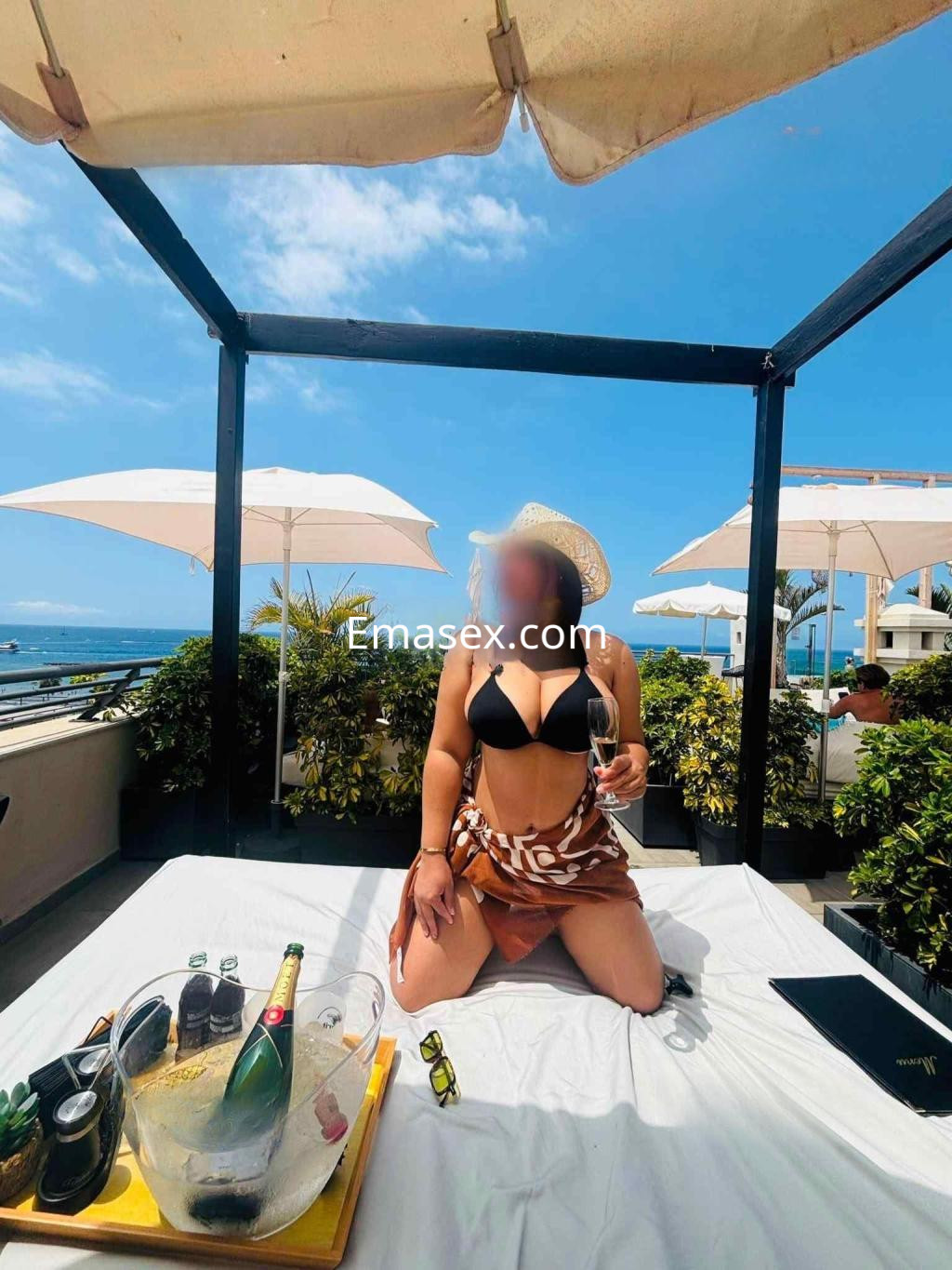 Puta posando en una terraza en Tenerife, con bikini negro, falda corta y sombrero, sosteniendo una bebida.