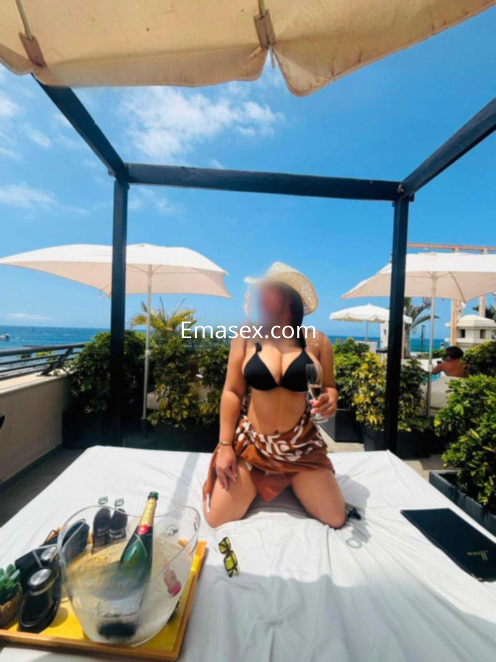 Puta en Tenerife, sentada en una cama al aire libre con un sombrero, luciendo un bikini negro y una falda ligera.