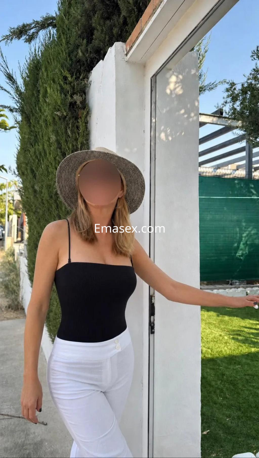 Puta en Zaragoza, con sombrero y blusa negra sin mangas, posando junto a una pared blanca y césped verde.