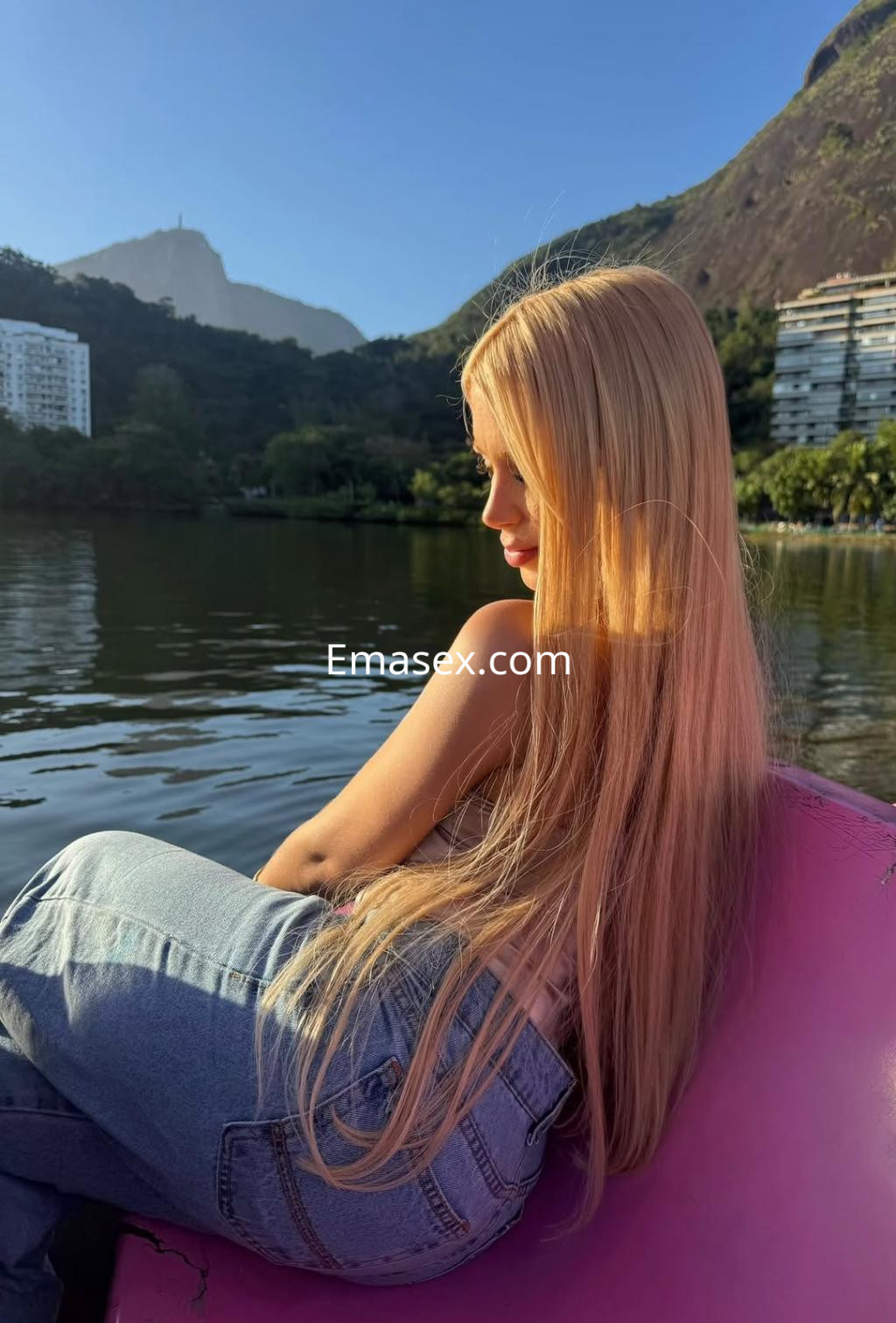Escort en Zaragoza con cabello largo y rubio, sentada de perfil junto a un lago, luciendo jeans claros.