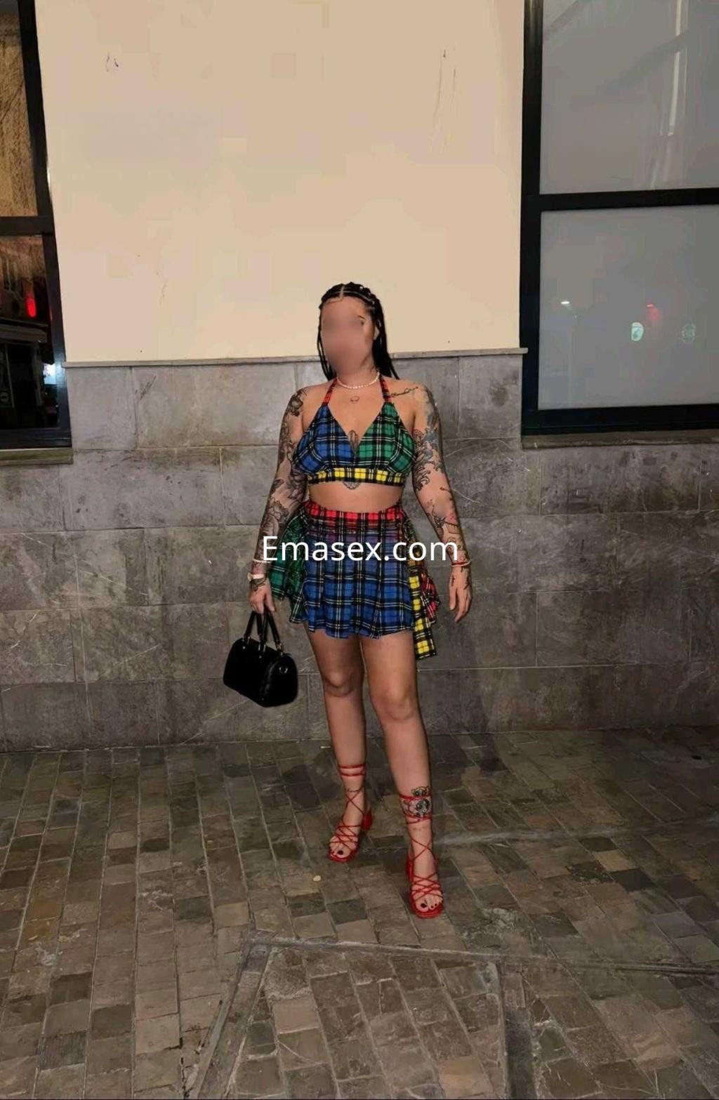 Escort en Almería con vestido de cuadros colorido, sandalias rojas y tatuajes visibles, posando en un entorno urbano.