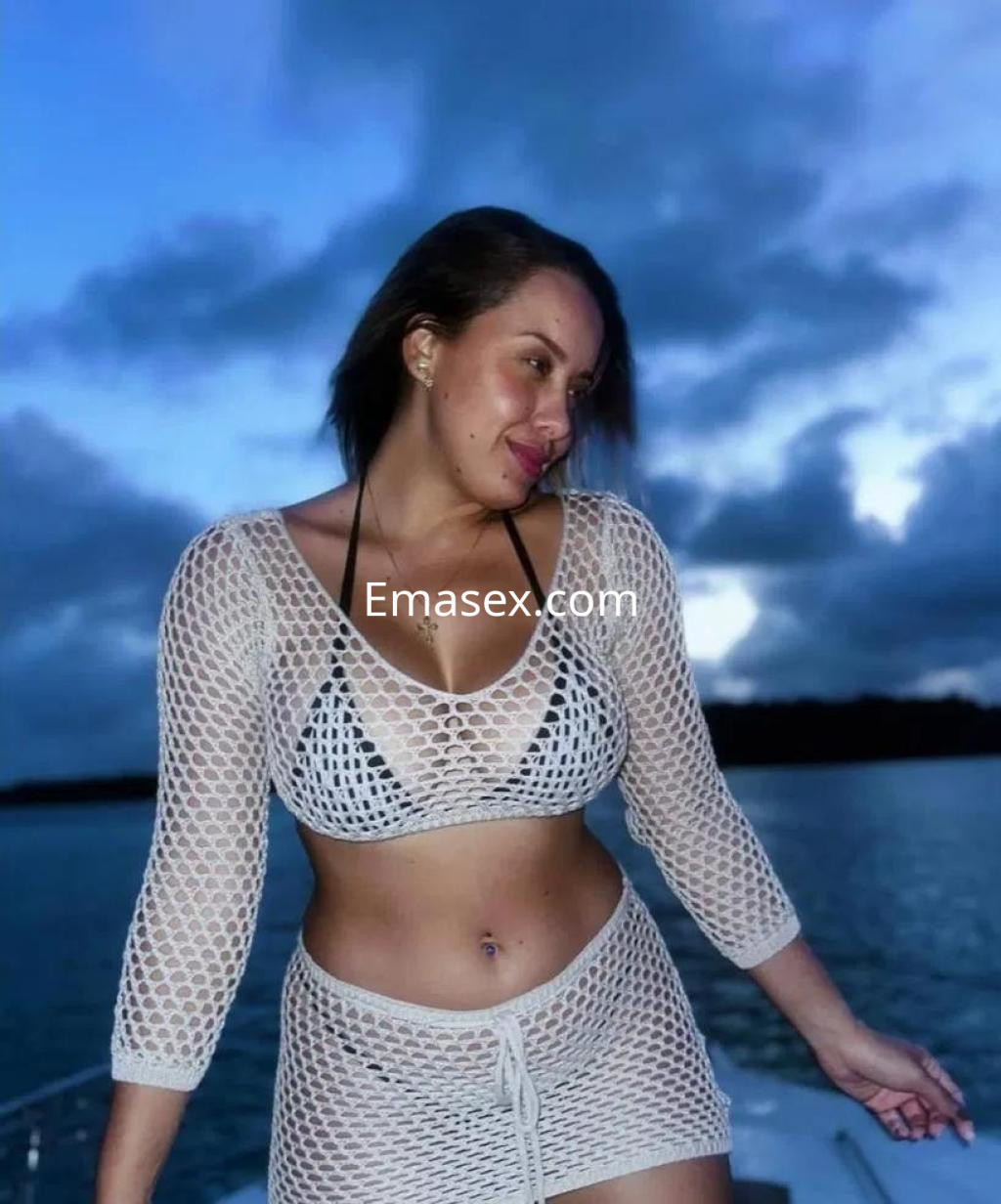 Escort en Vitoria luciendo un bikini negro y una blusa de malla blanca, posando junto al mar al atardecer.