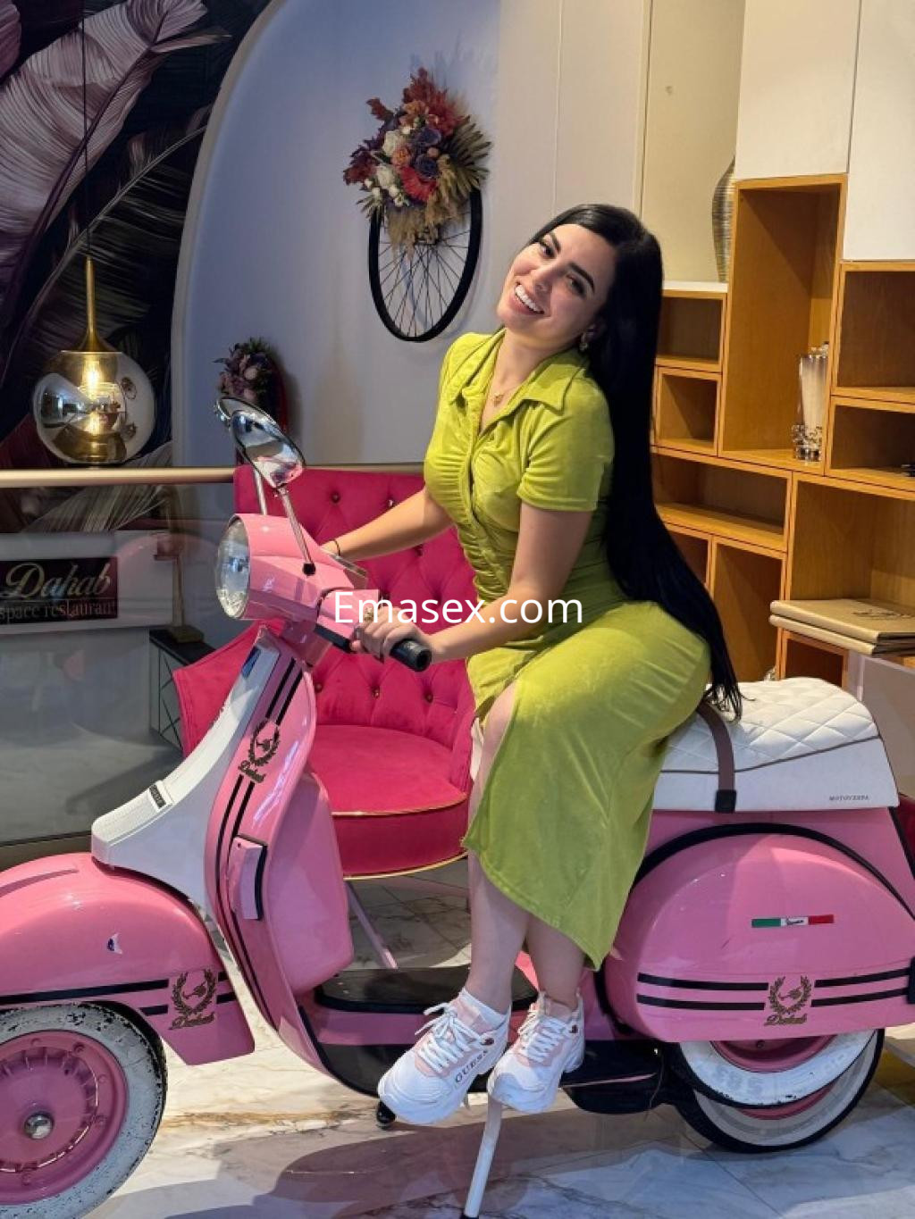 Escort posando sonriente sobre una scooter rosa, con vestido verde y zapatillas blancas en un entorno moderno en Huelva.